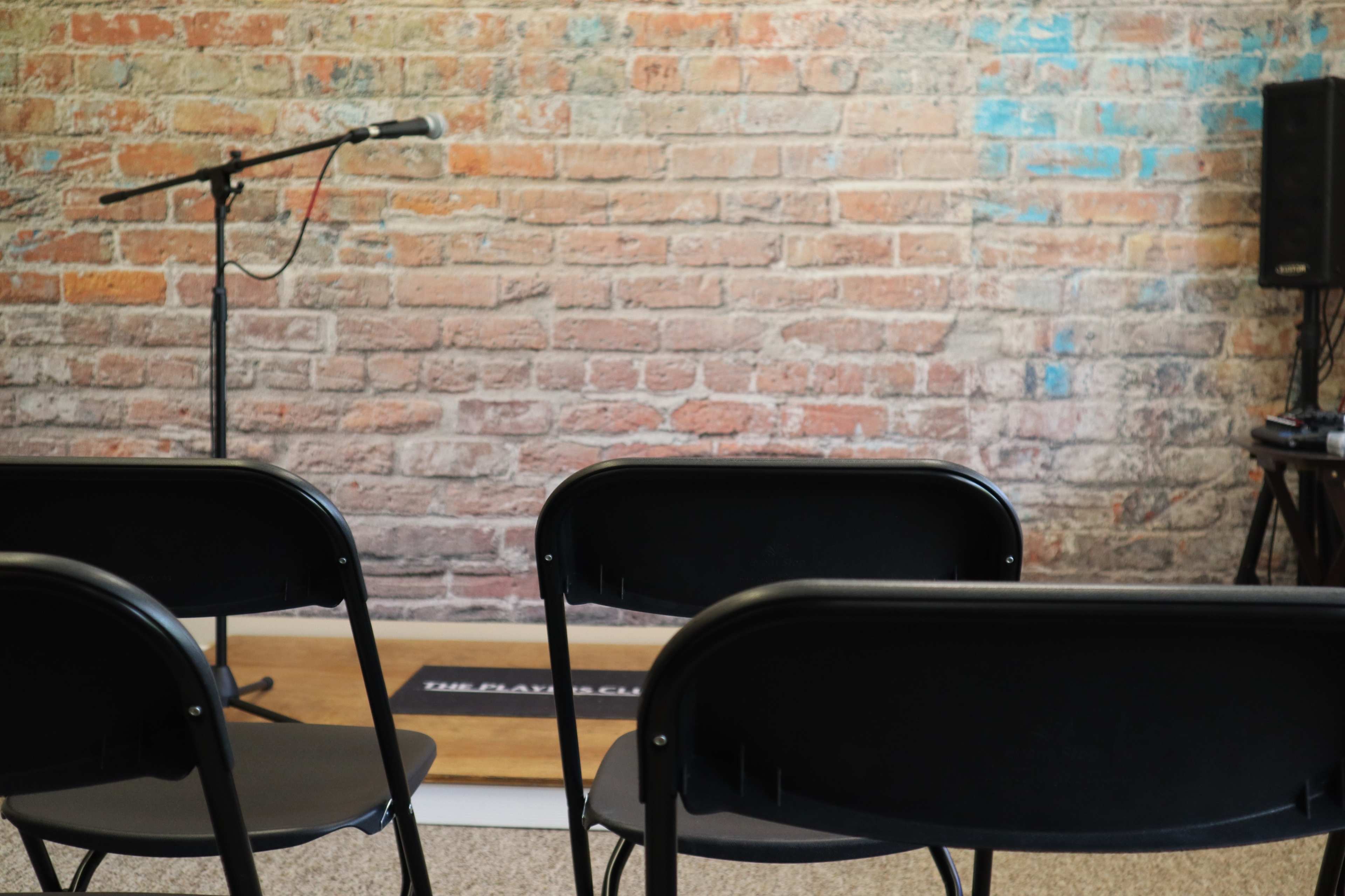 A row of black folding chairs faces a microphone on a stand in front of a brick wall.