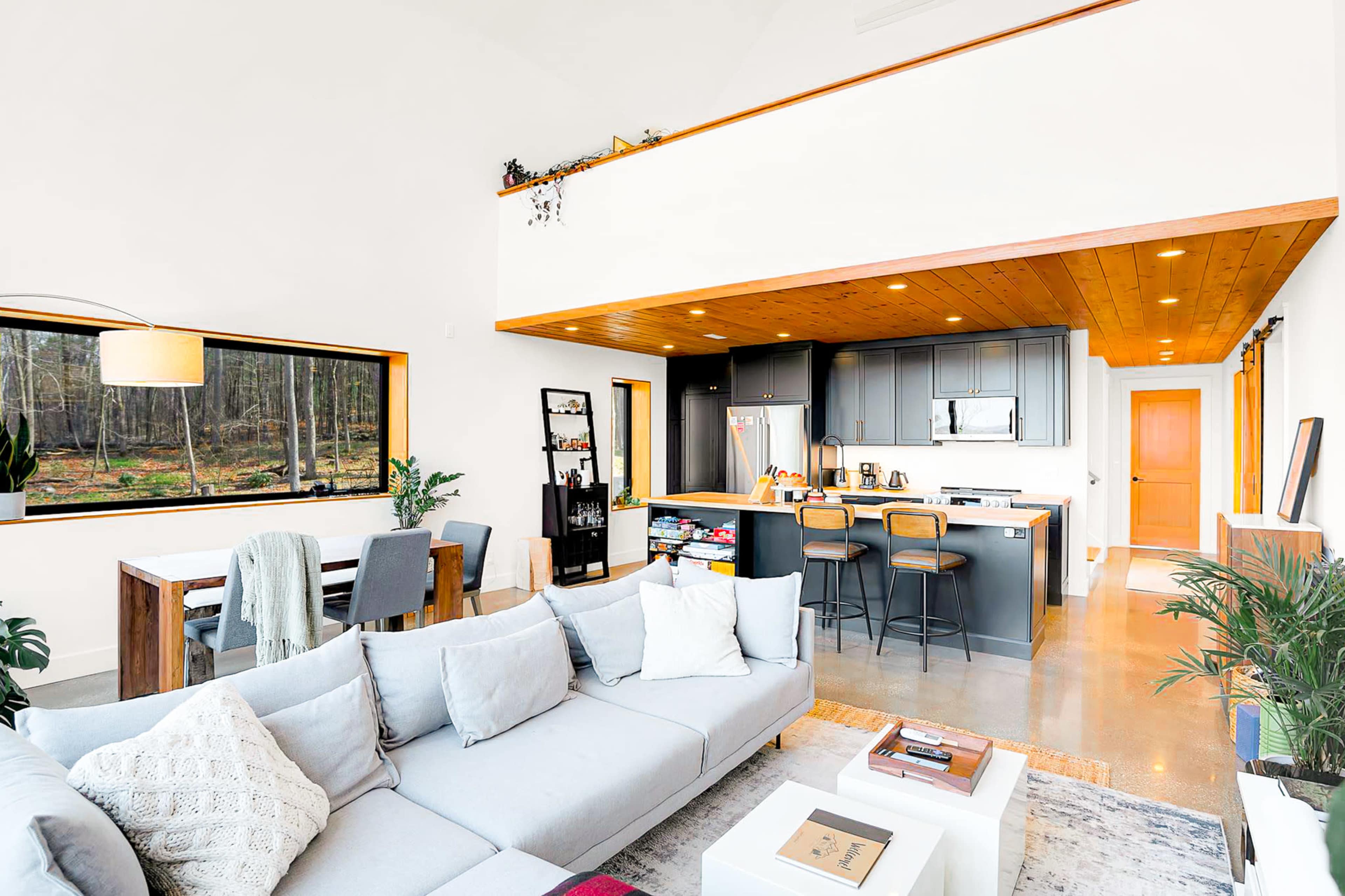 A modern living space featuring a light grey sectional sofa, a dining area with a table and chairs, and a kitchen with dark grey cabinetry and stainless steel appliances, all illuminated by natural light from large windows.