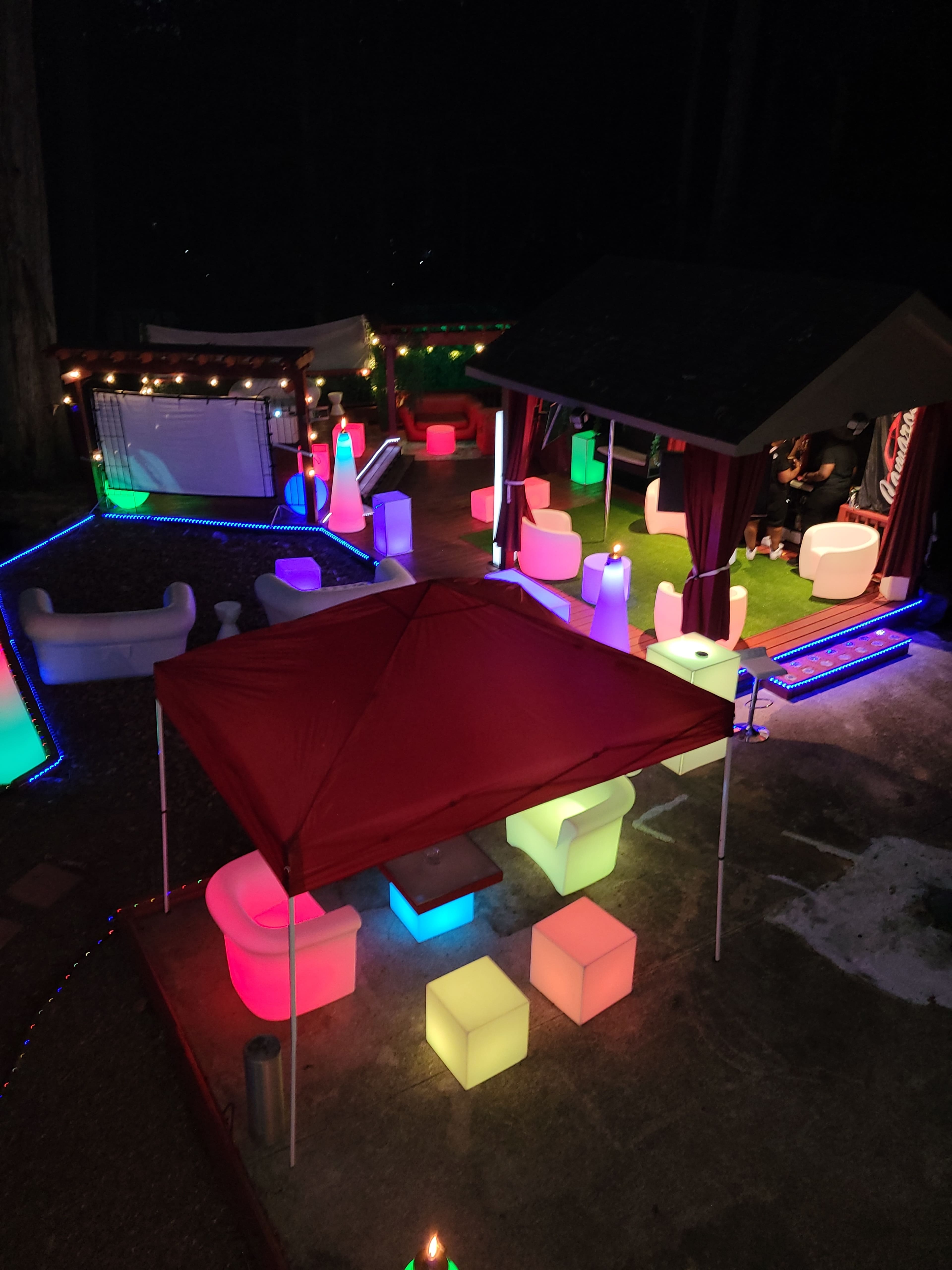 The scene features a nighttime outdoor venue illuminated by colorful lighting, with illuminated furniture and a red canopy in the center.