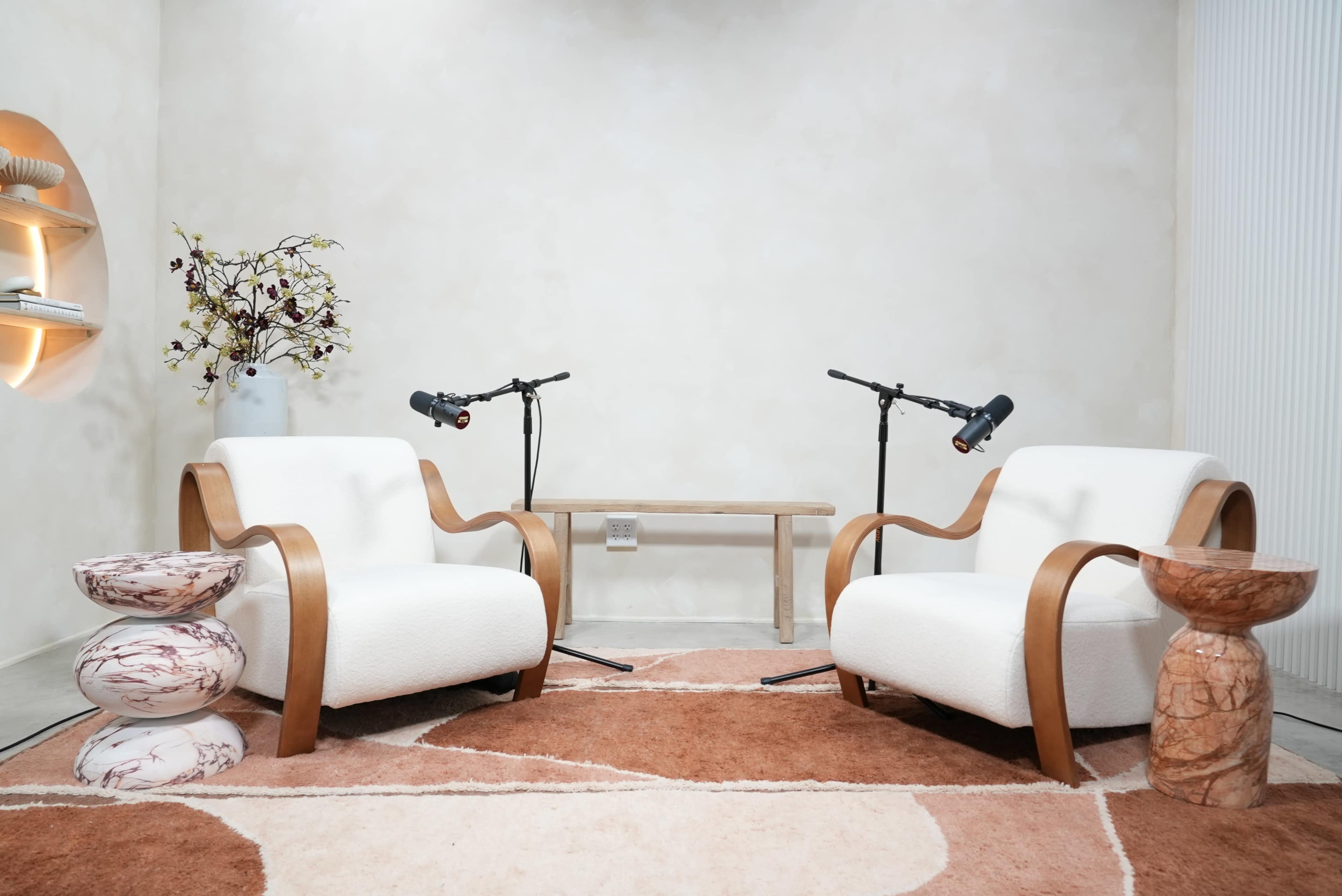 The image shows a minimalist interior with two upholstered chairs, a wooden table, and decorative elements, including a rug and small side tables.