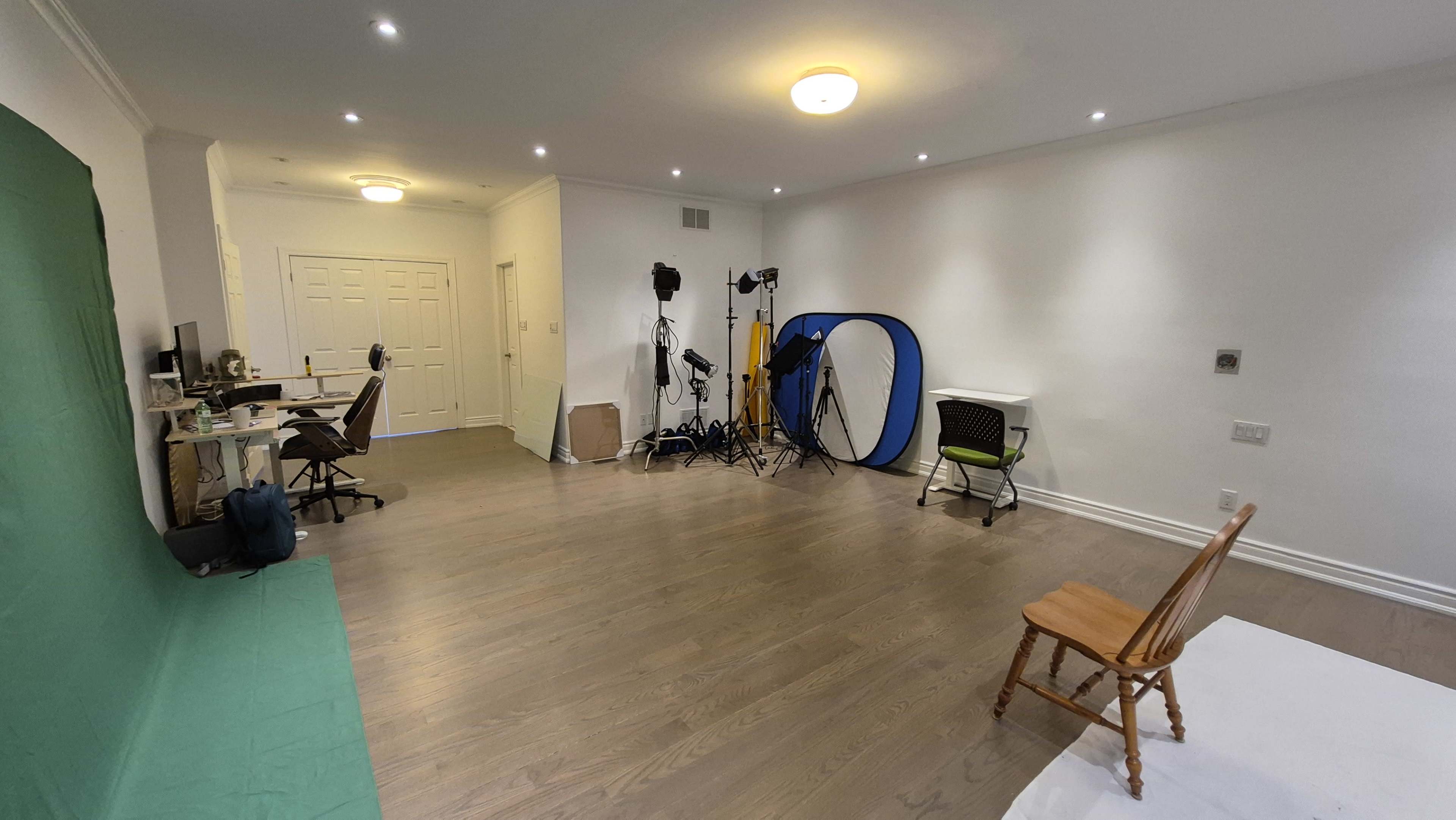 The room features a green screen backdrop, a desk with a computer, various lighting equipment, and a single wooden chair.