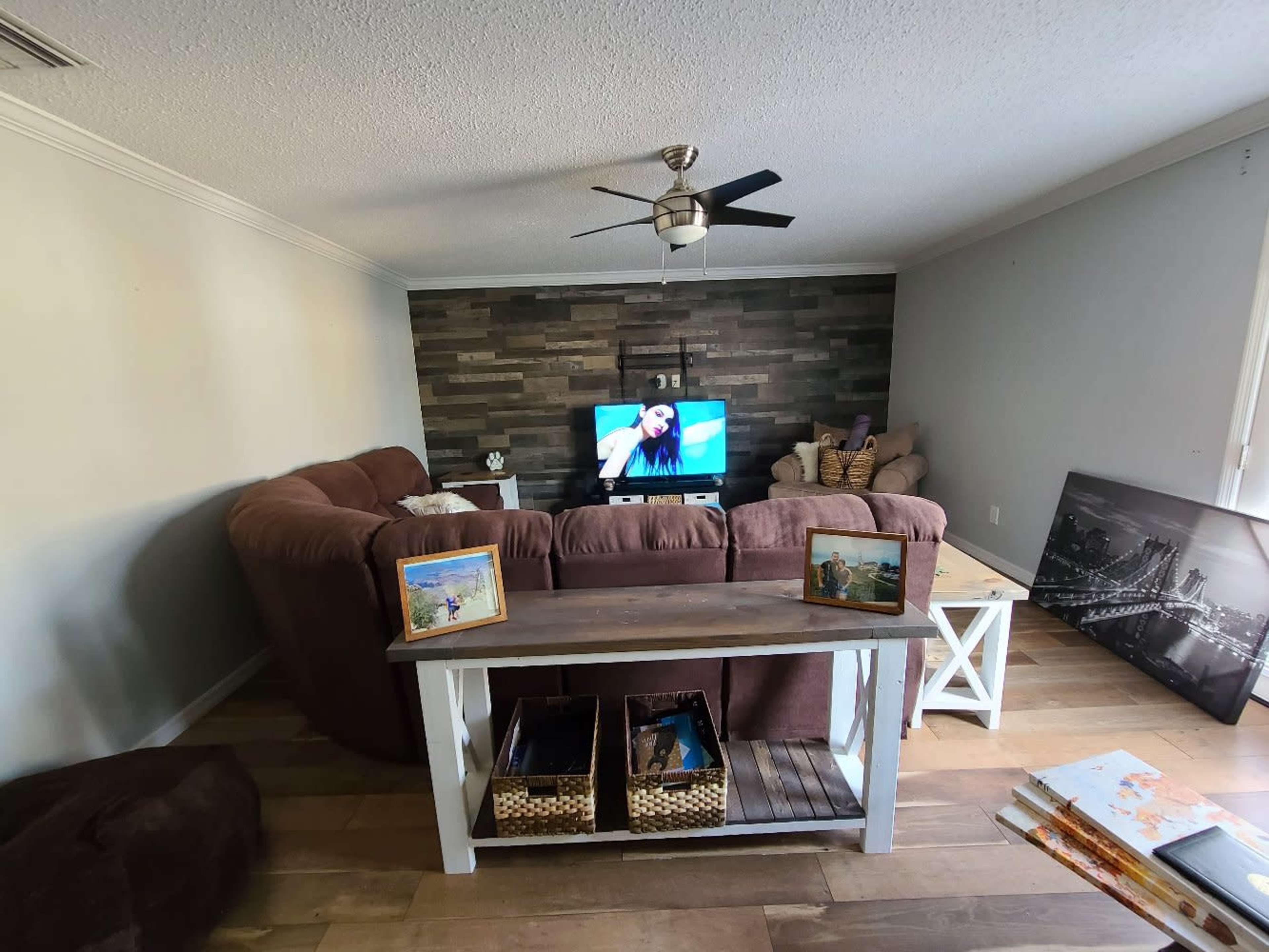 A living room features a large brown sectional sofa facing a television on a stone-paneled wall, with a wooden coffee table displaying framed photos and decorative storage baskets.