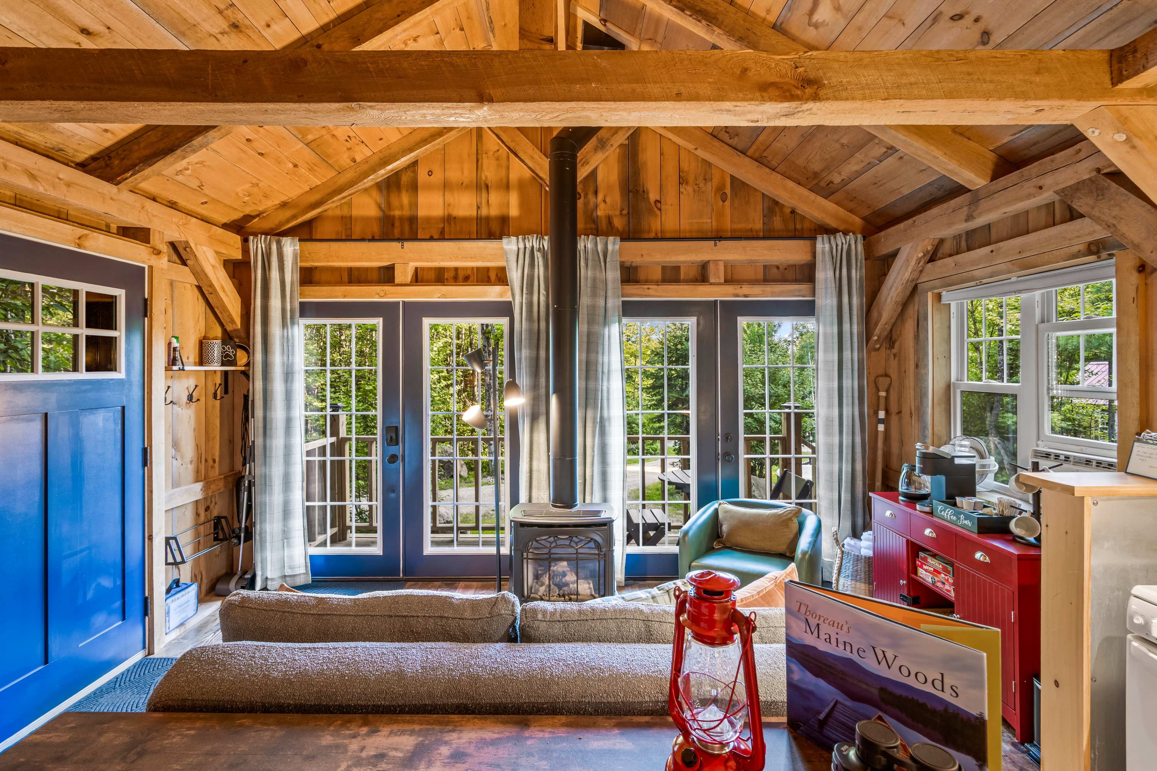 The interior of a wooden cabin features a cozy living space with a stove, large windows, and a combination of rustic decor and furniture.