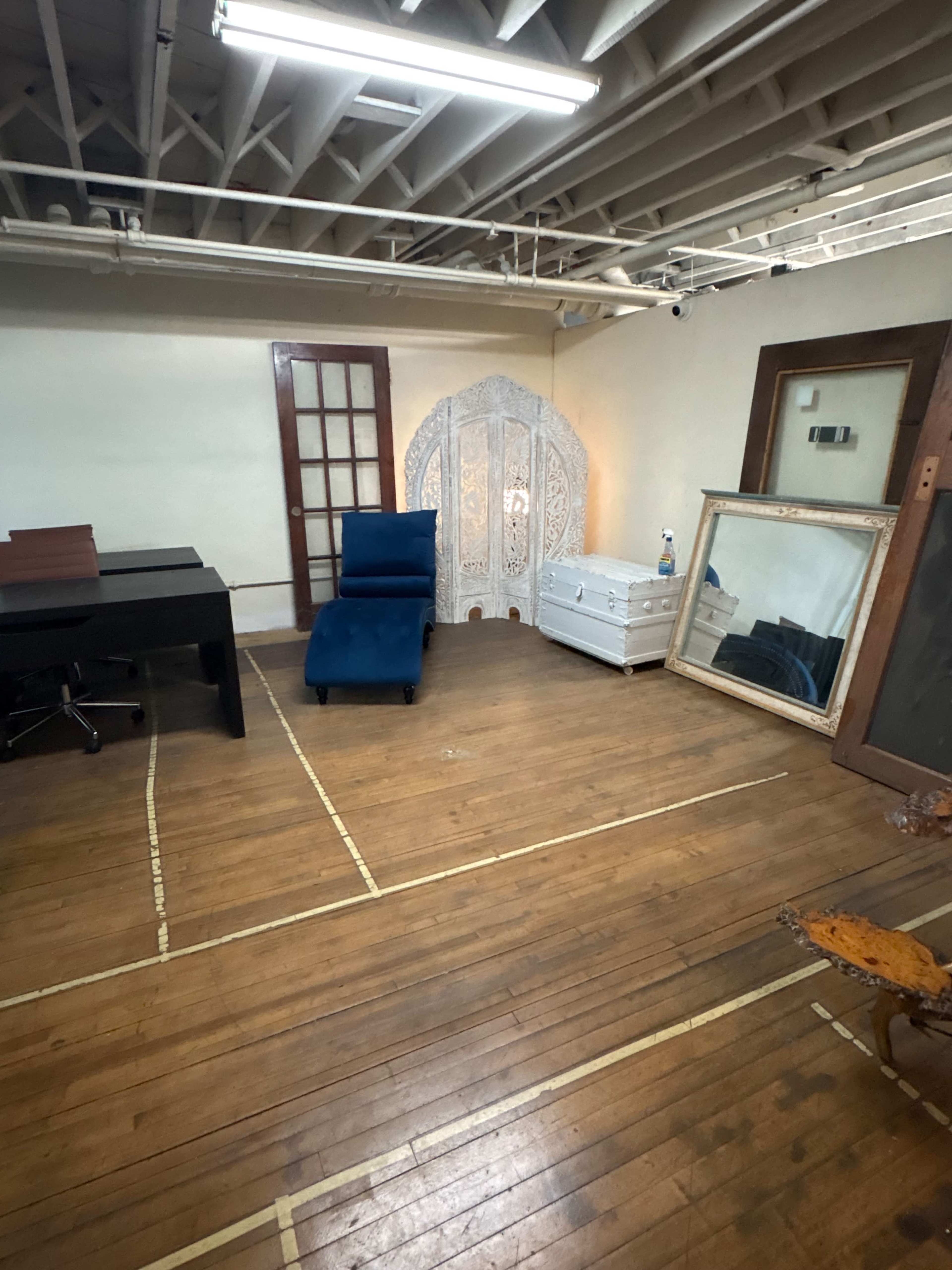 The image shows a spacious, empty room with wooden flooring, featuring a blue chair, a large mirror, and an intricately carved white screen against one wall.