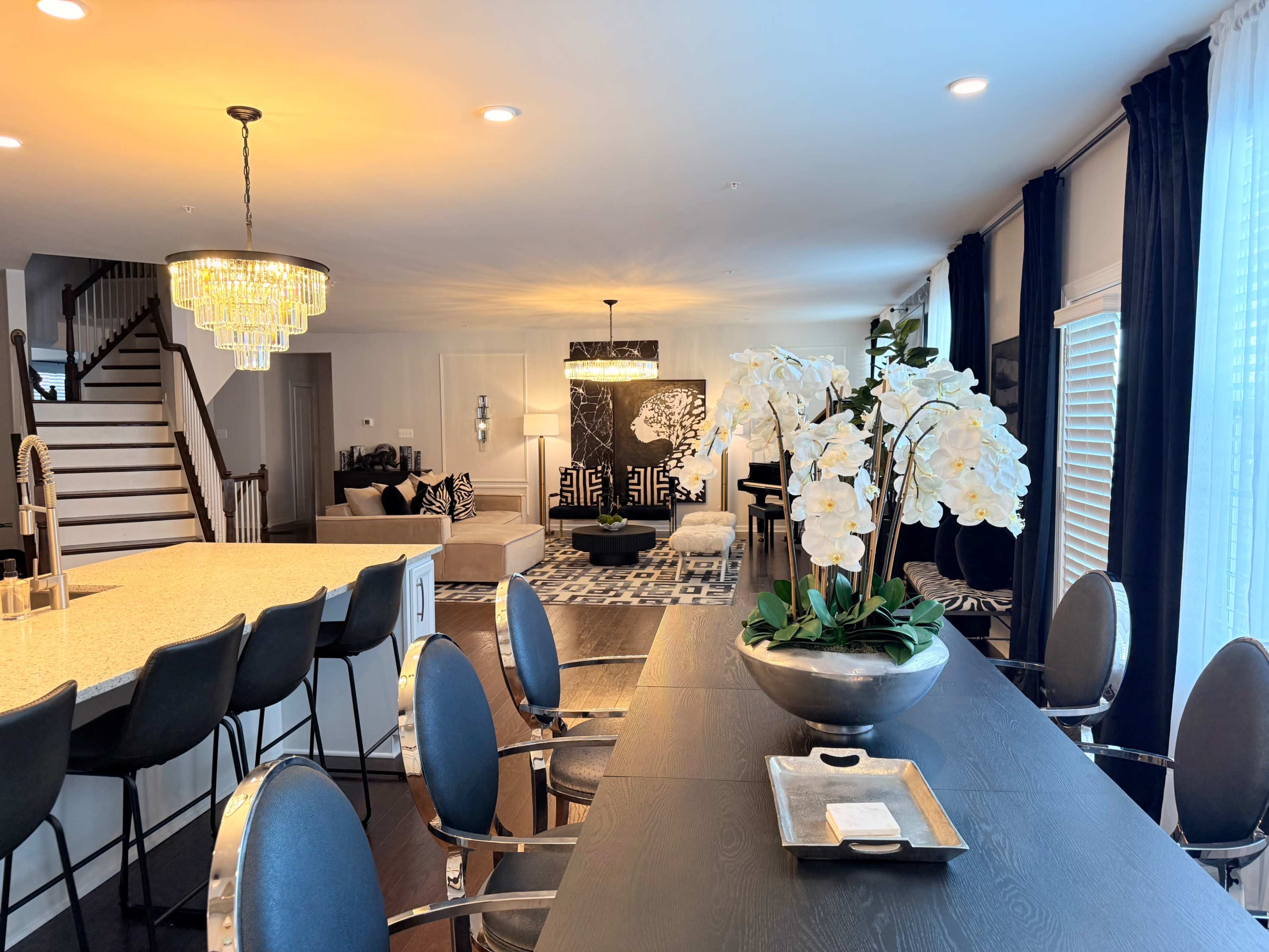 The image shows a modern living area and kitchen with a large island, elegant lighting fixtures, and decorative plants arranged on a dining table.