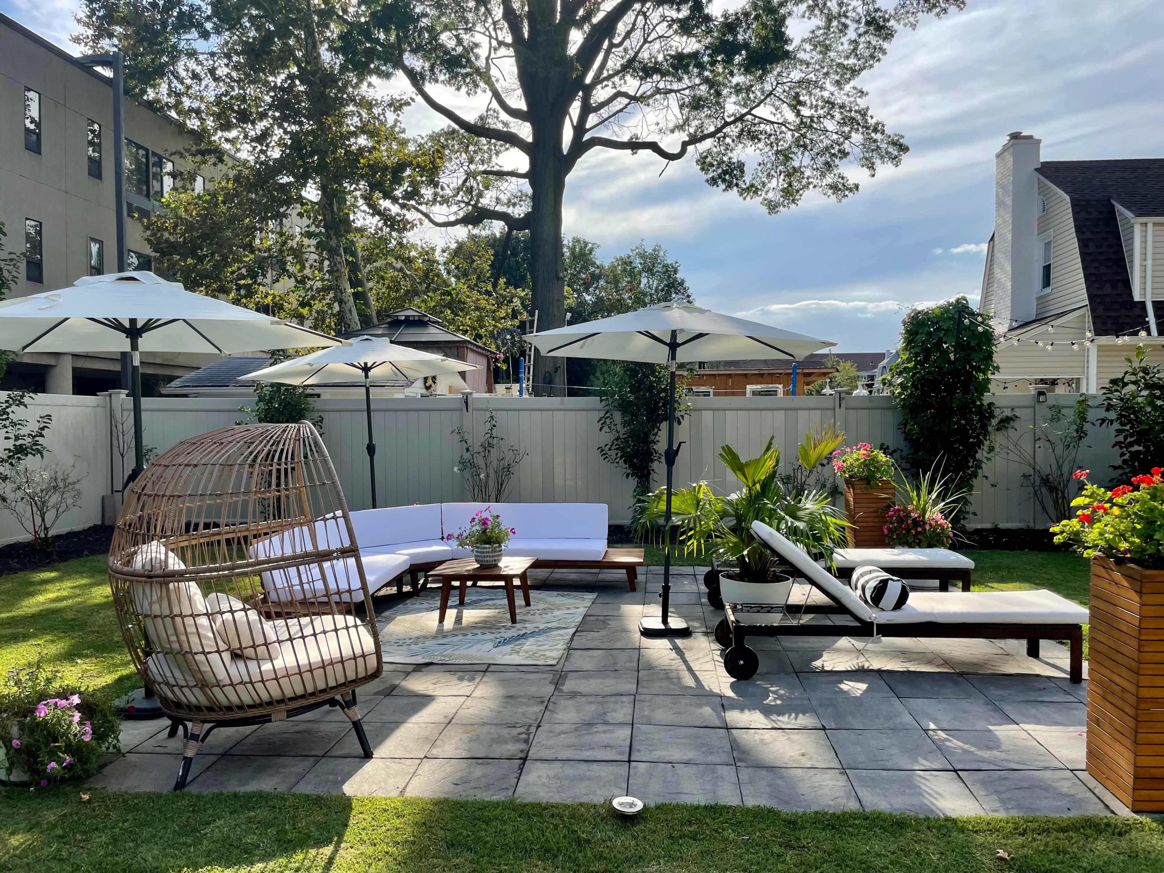 A neatly arranged outdoor living area featuring a circular wicker chair, a white sectional sofa, and two lounge chairs under large umbrellas, surrounded by potted plants and a paved patio.