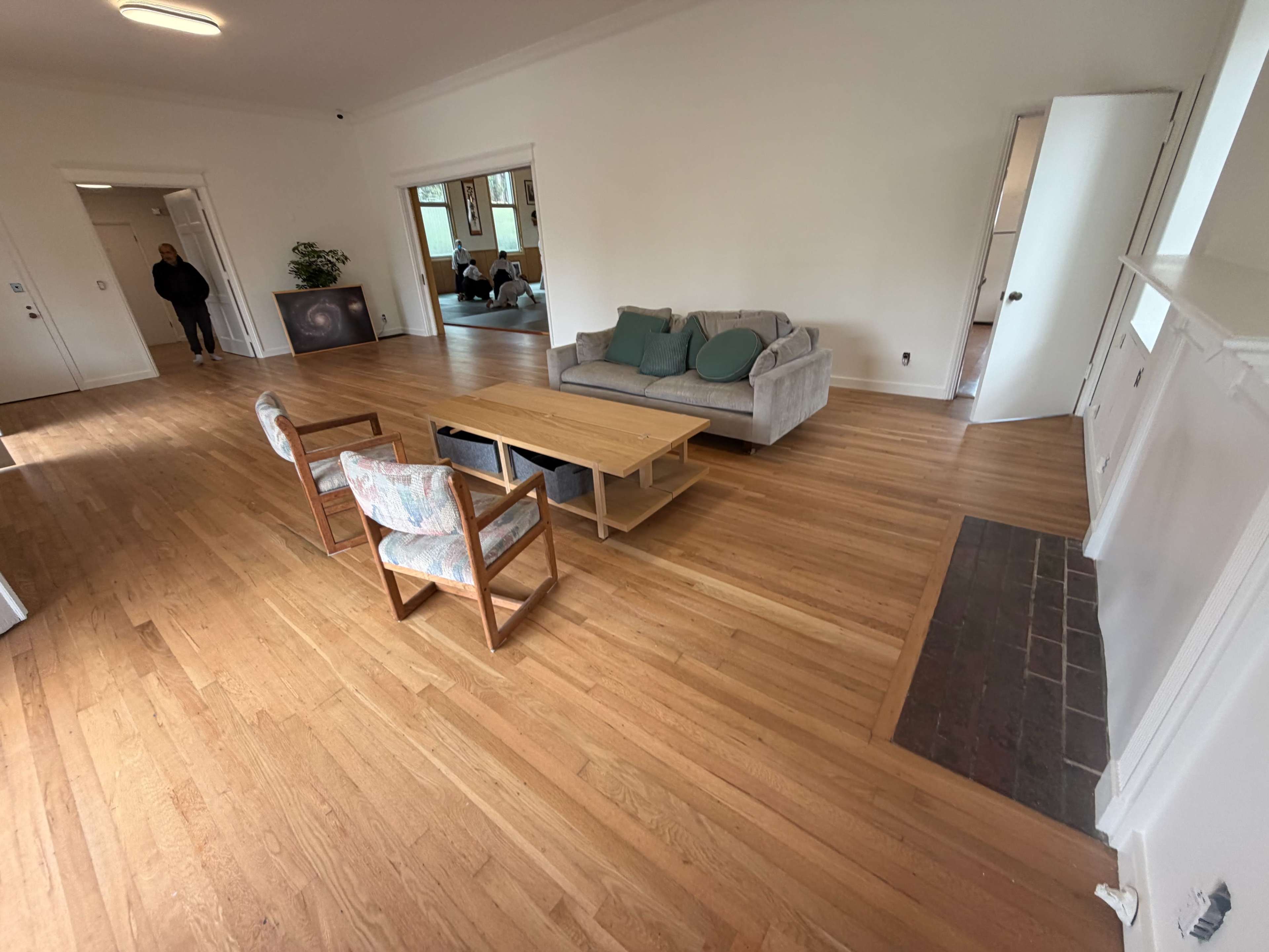 The image shows a spacious, empty room with wooden flooring, featuring a gray couch and a coffee table in the center, surrounded by two chairs.