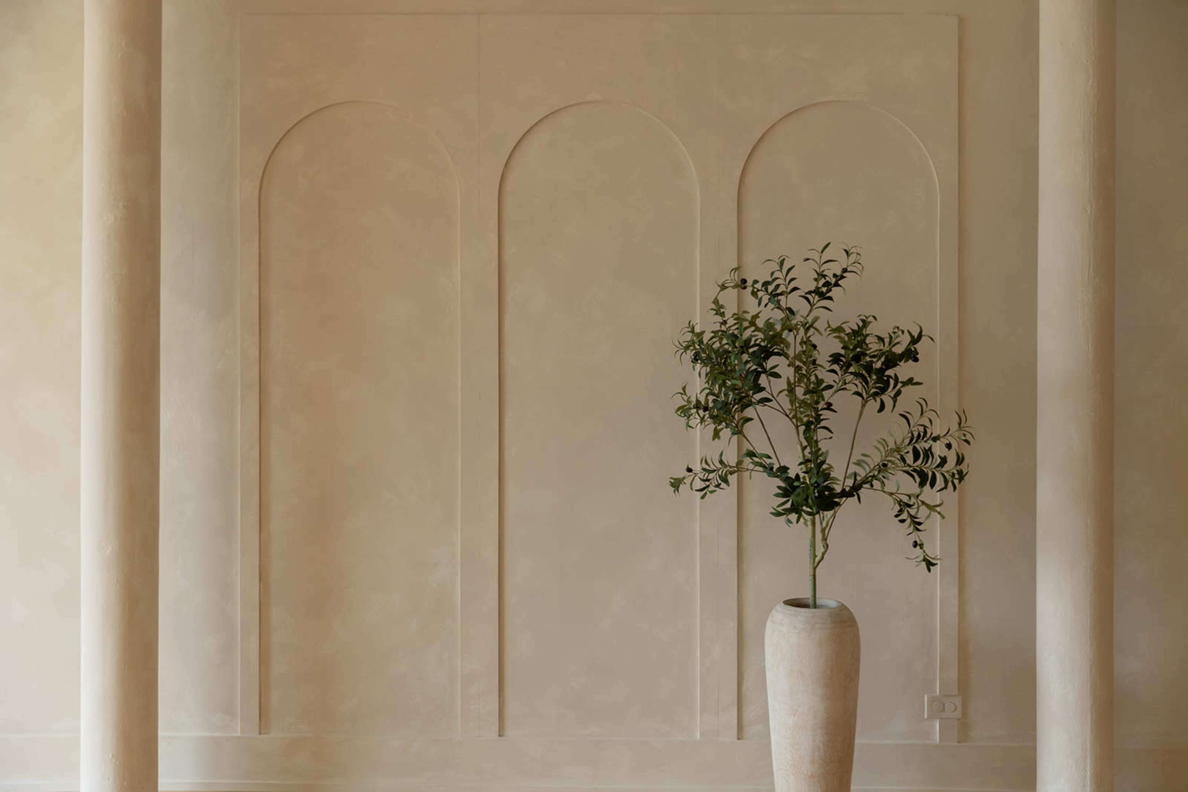A tall, neutral-colored vase with a leafy plant stands against a textured wall featuring three arched niches.
