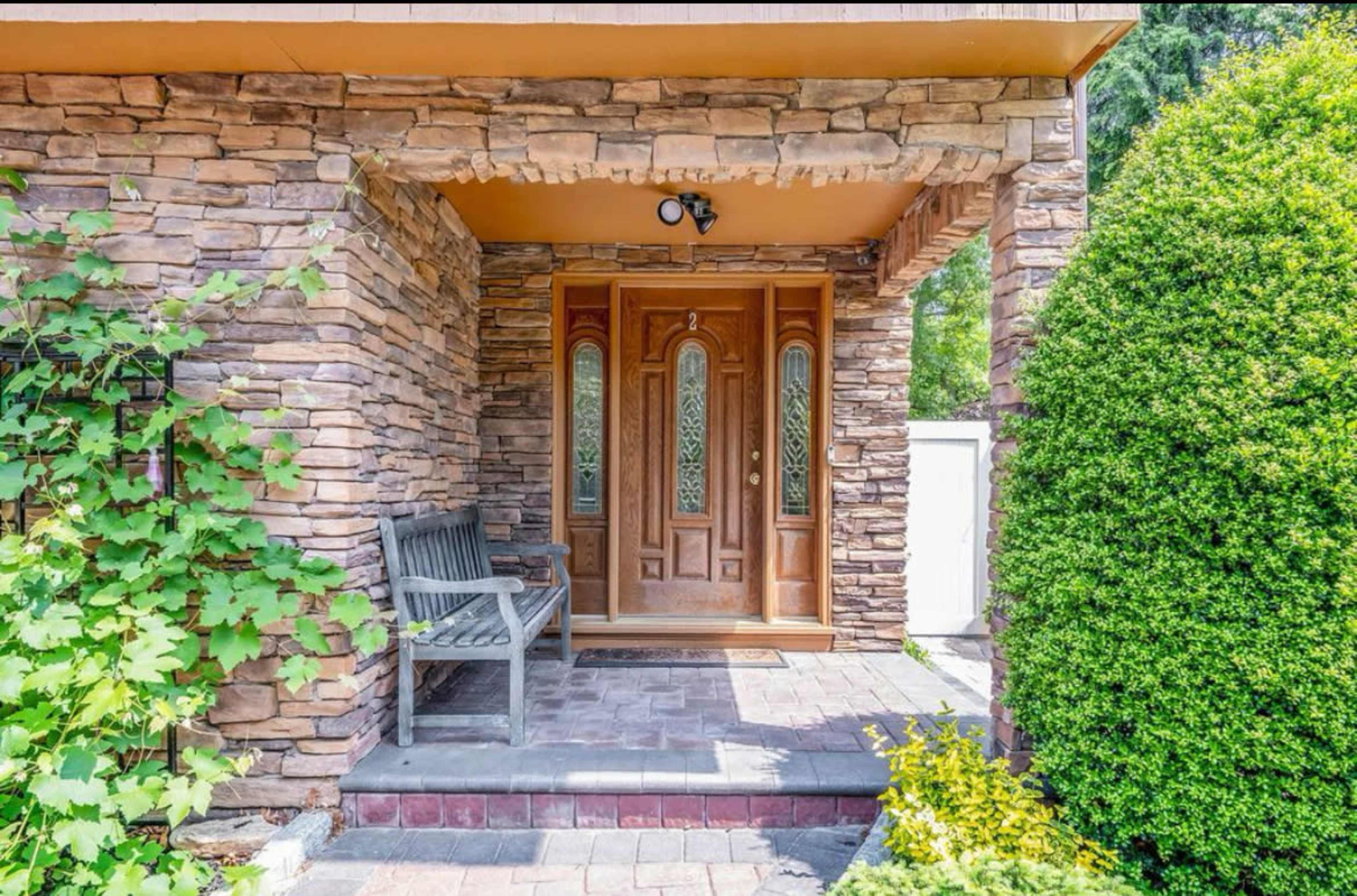 The image shows a stone-clad entrance with a wooden door framed by decorative windows and flanked by a bench and lush greenery.