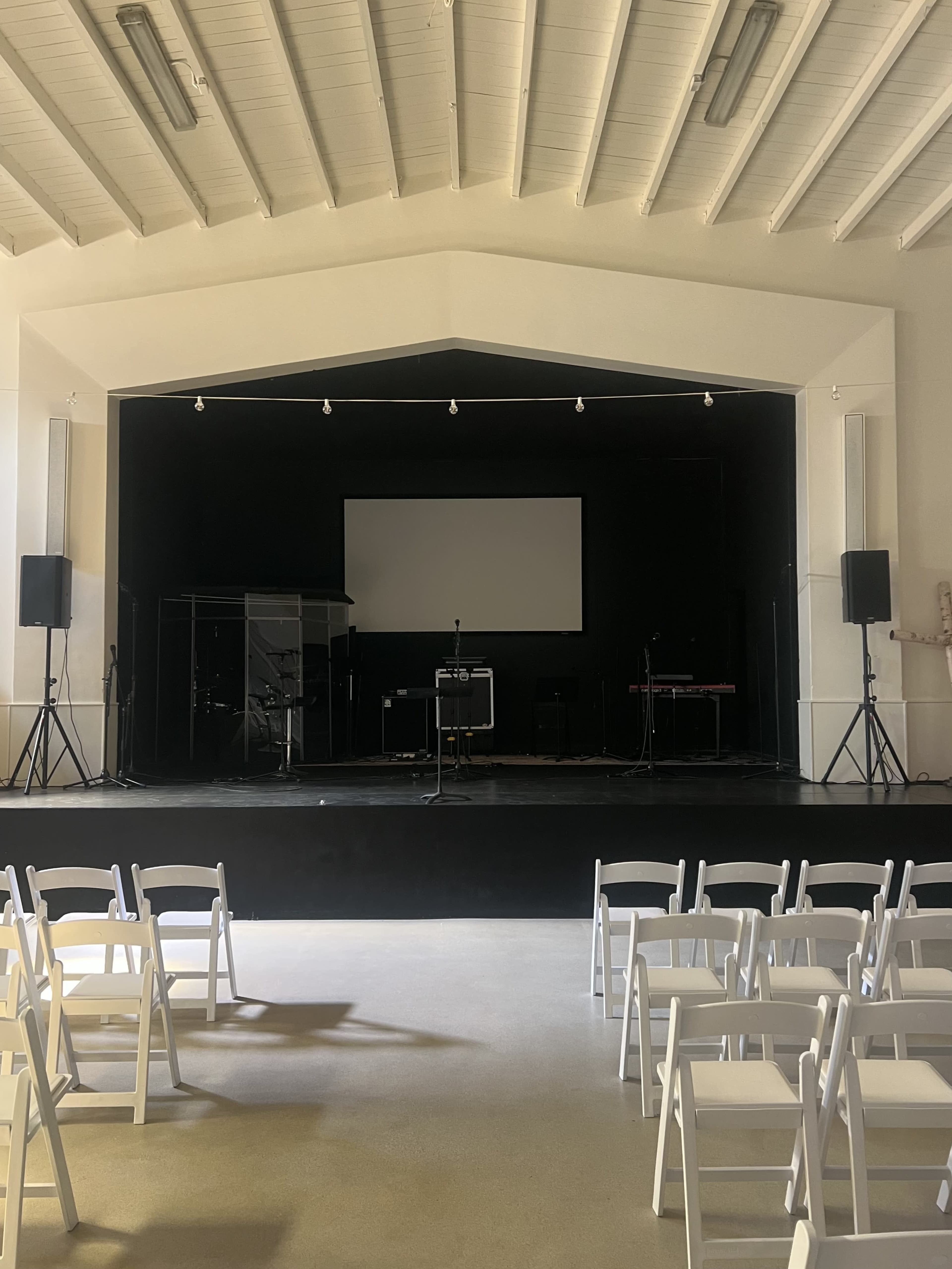 The image shows a stage area in a hall, featuring a blank screen, musical instruments, and rows of white folding chairs facing the stage.