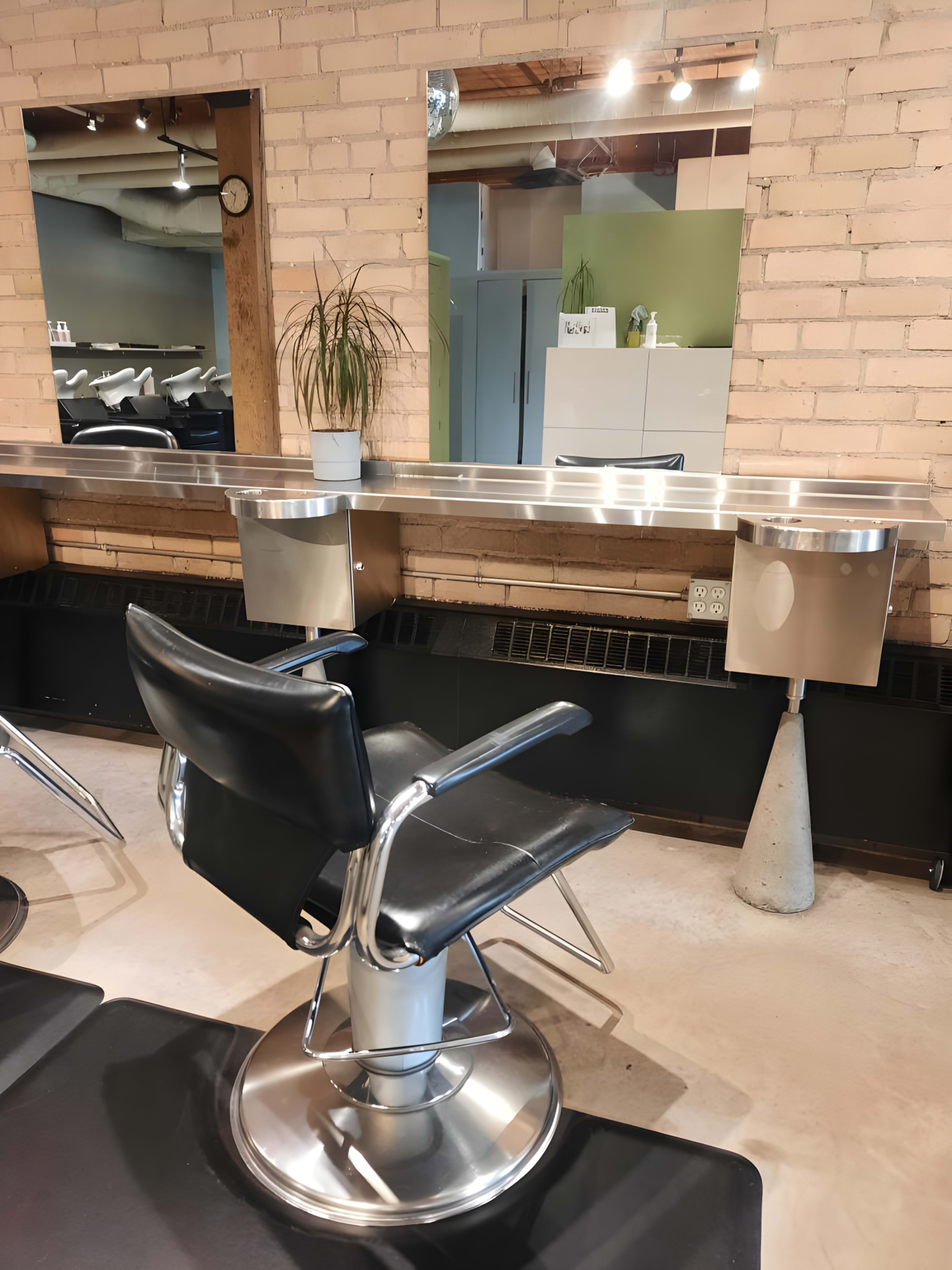A black salon chair is positioned in front of a mirror mounted on a brick wall, with a potted plant on the counter and hair washing stations visible in the background.