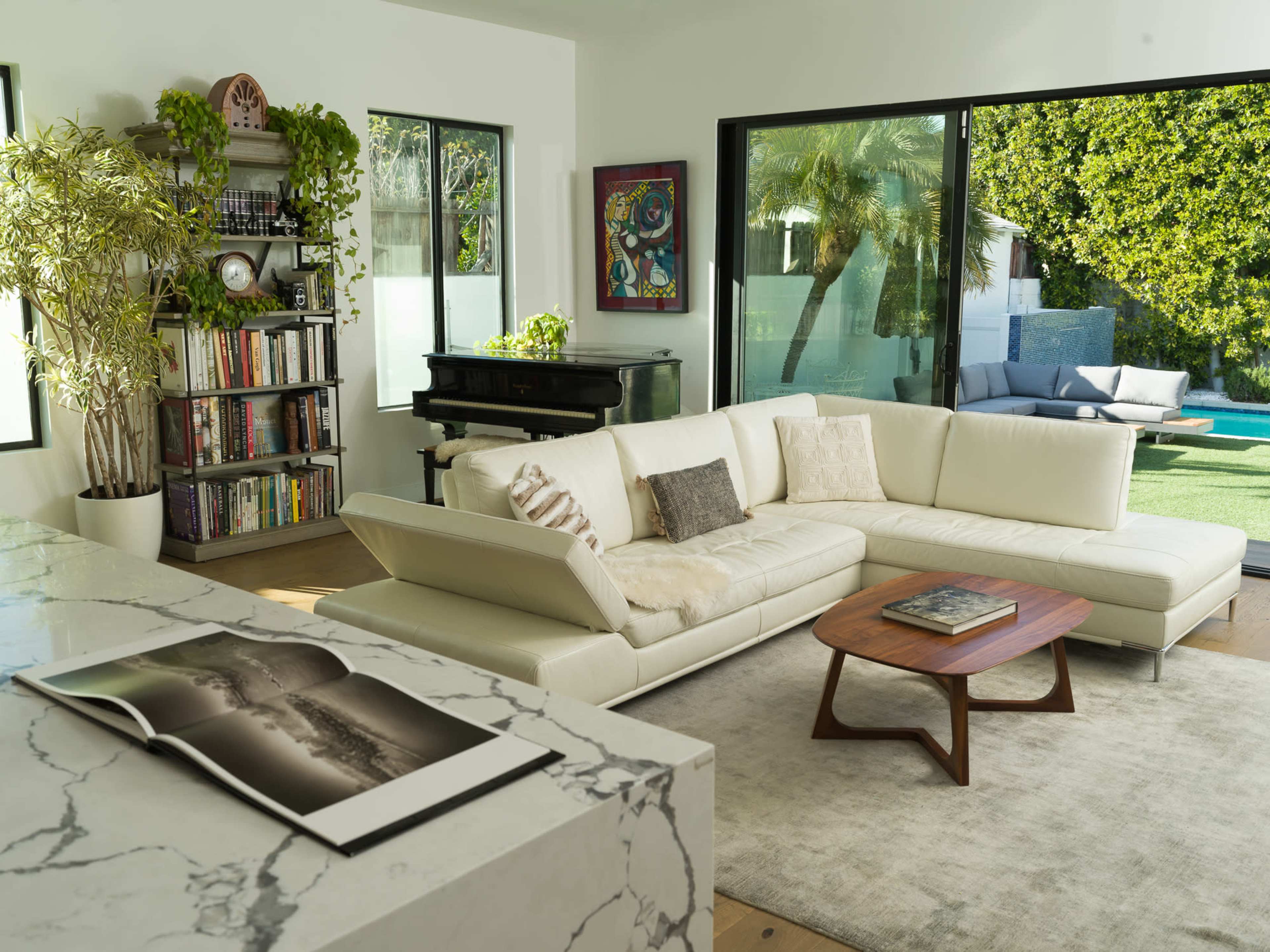 A modern living room features a white sectional sofa, a coffee table, a marble island, a bookshelf, and large glass windows overlooking a yard with a pool.