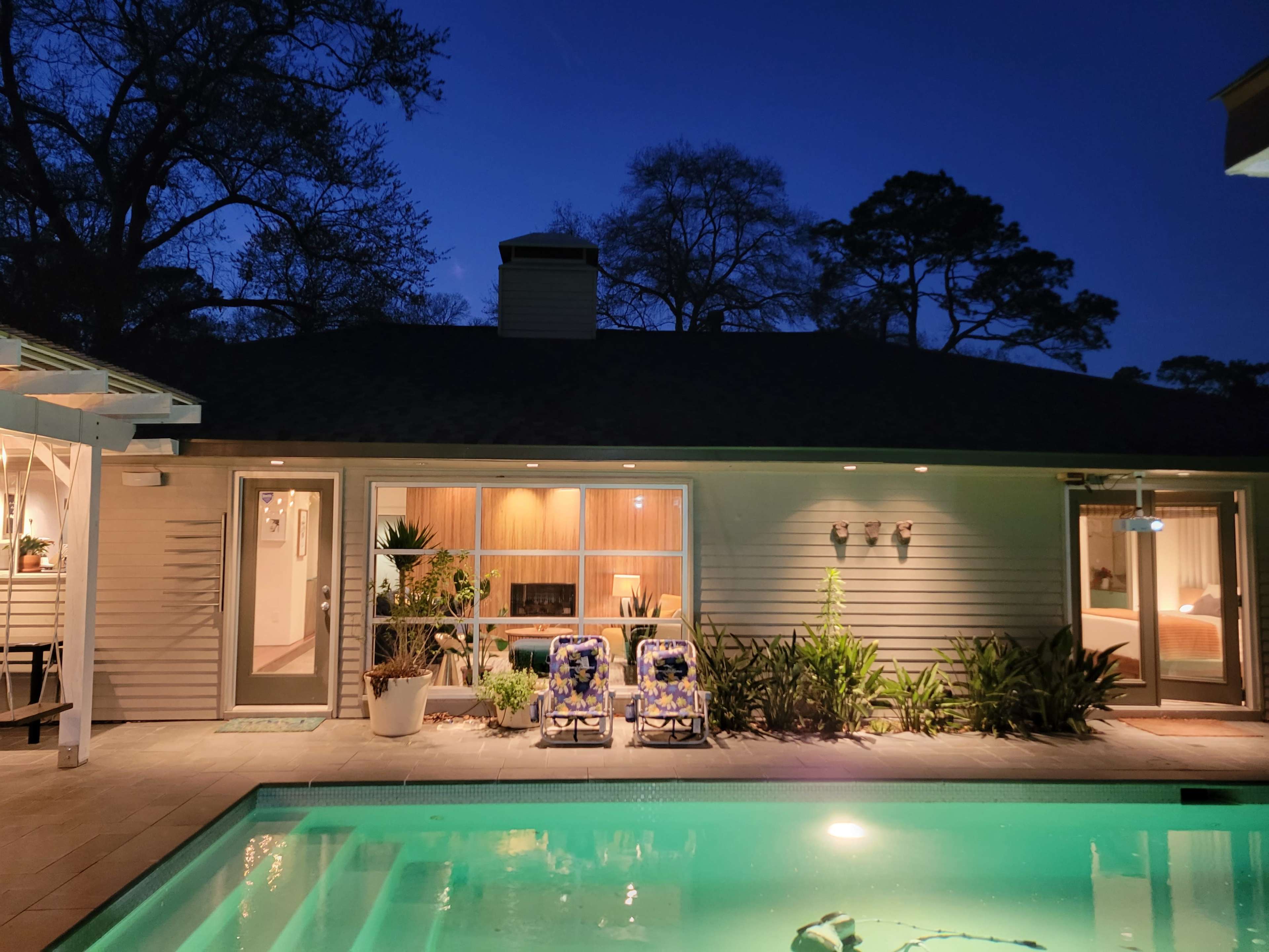 A modern home with large windows overlooks a pool, illuminated in the evening light, surrounded by lush greenery.