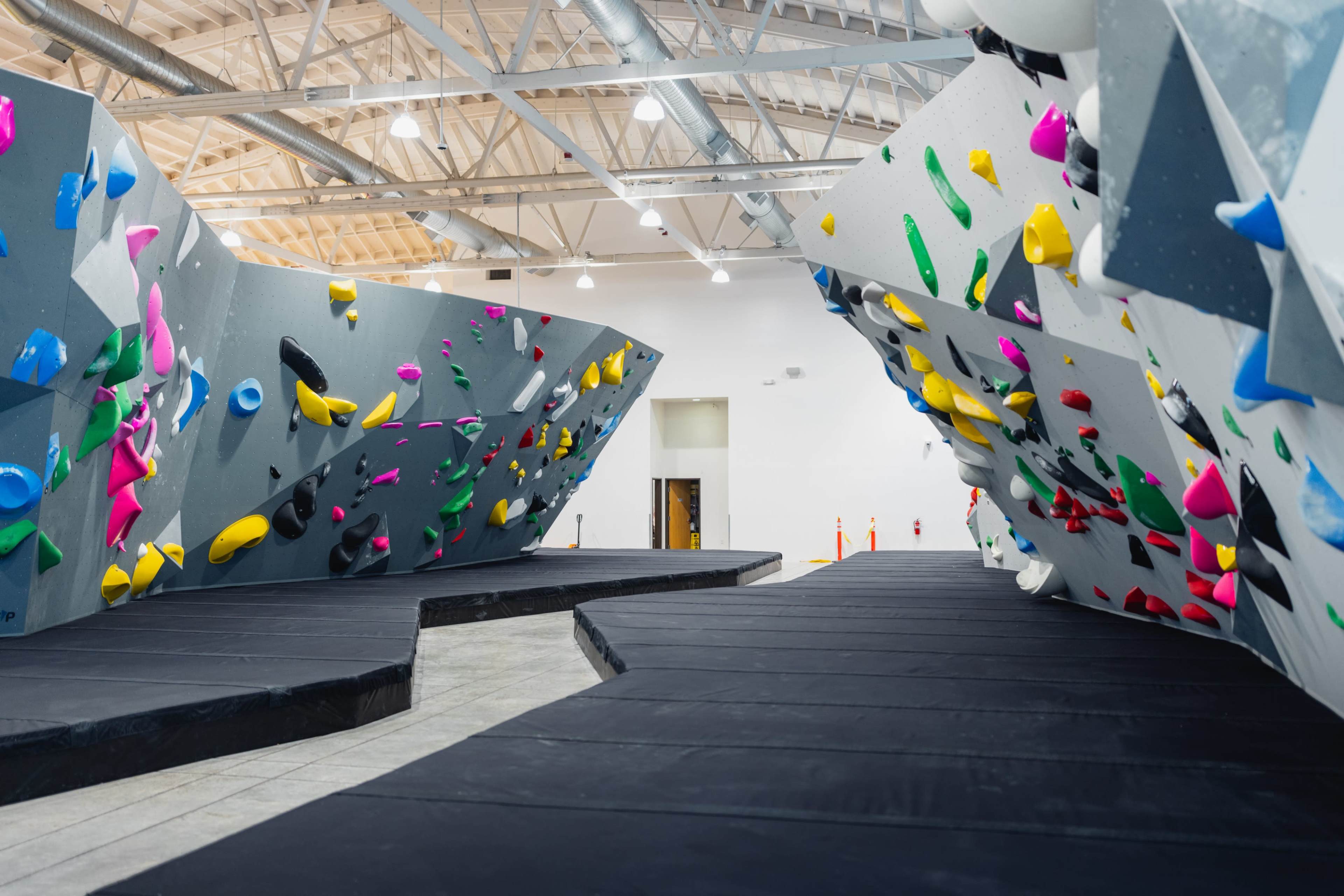 The image shows an indoor bouldering gym featuring multiple climbing walls with various colored holds and a padded floor for safety.