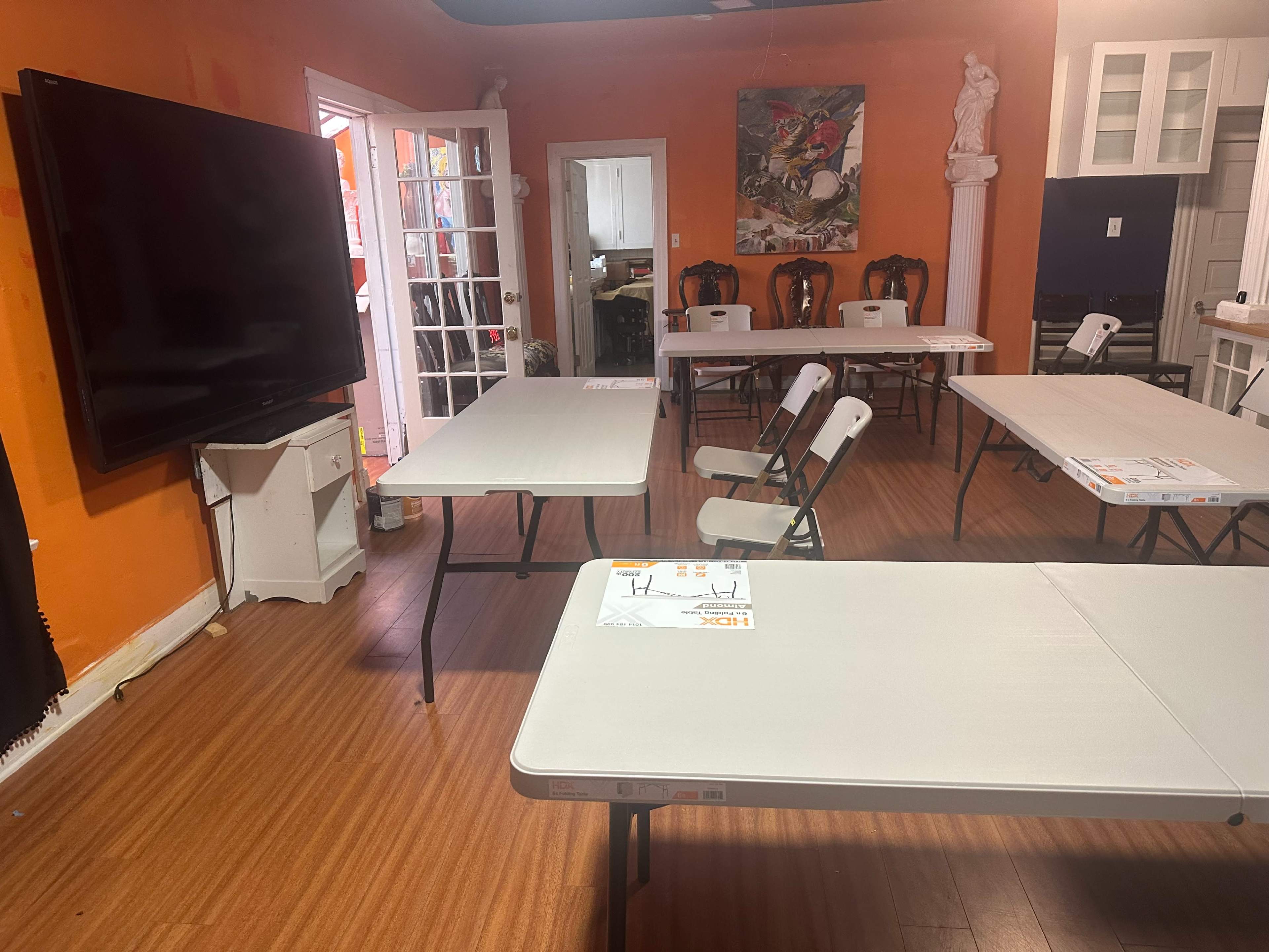 The image shows an orange-walled room with several folding tables and chairs arranged around a large television.