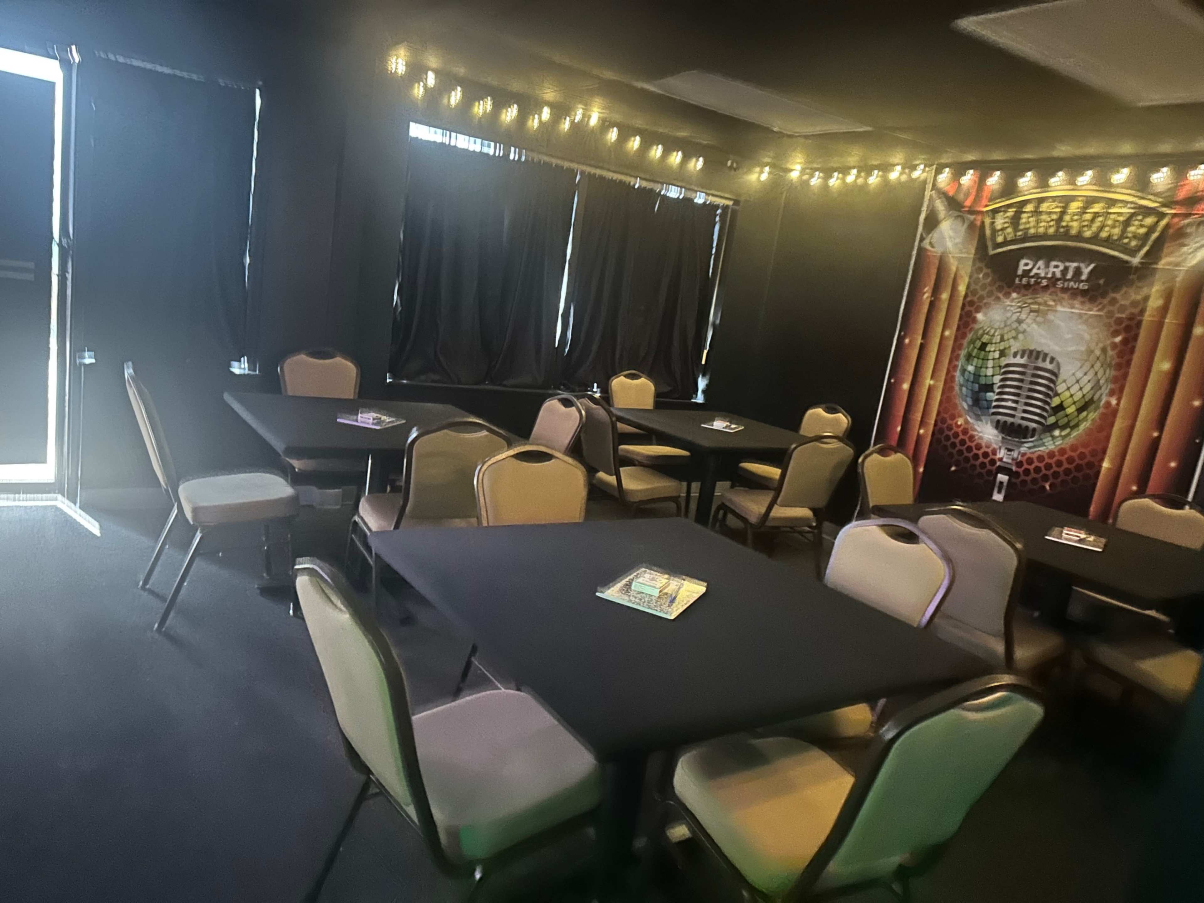 The image shows an indoor venue with several round tables and chairs, dim lighting, and a decorated wall featuring a microphone motif.
