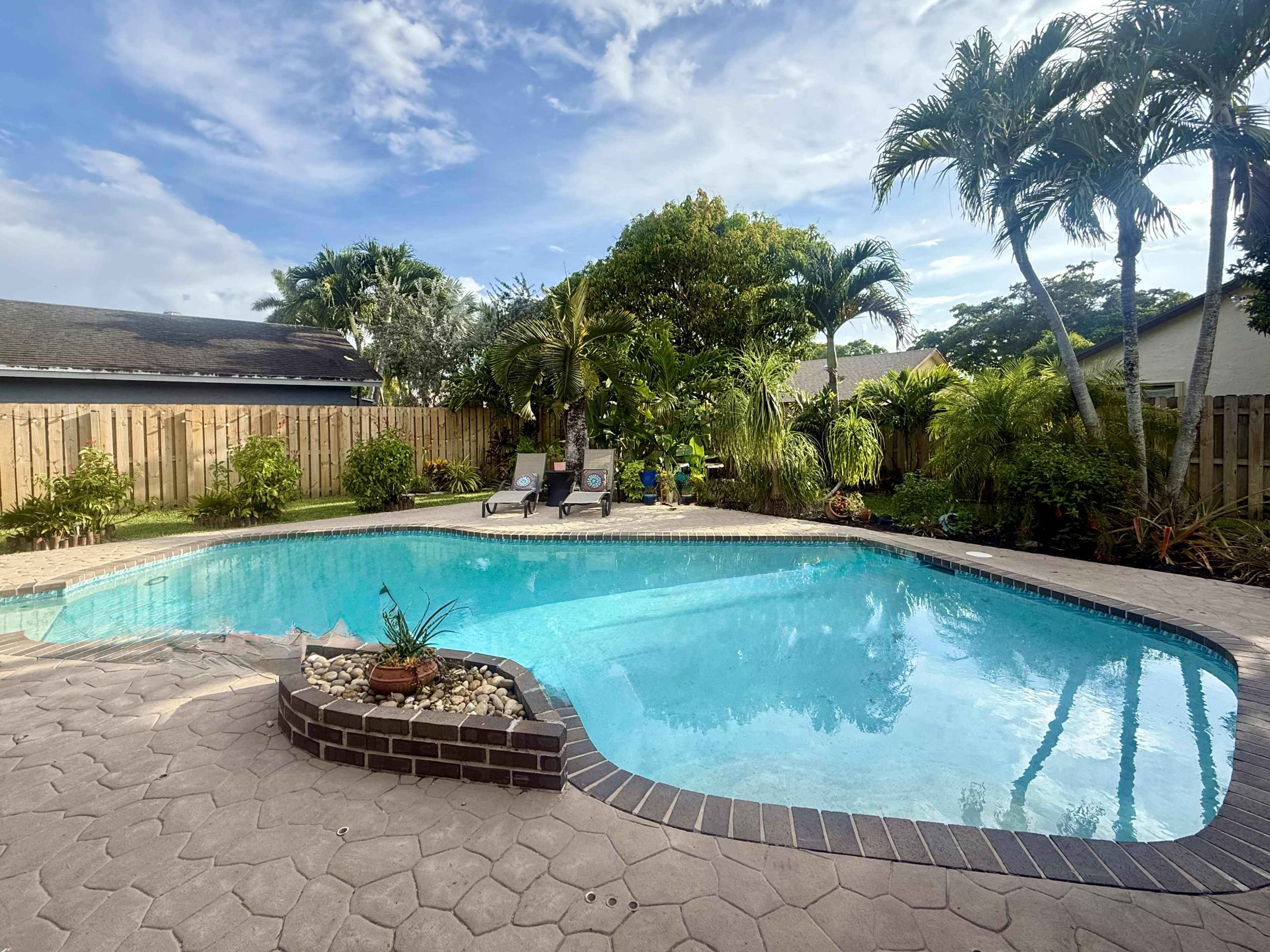 The scene shows a clear blue swimming pool surrounded by tropical plants and palm trees, with two lounge chairs placed on the patio.