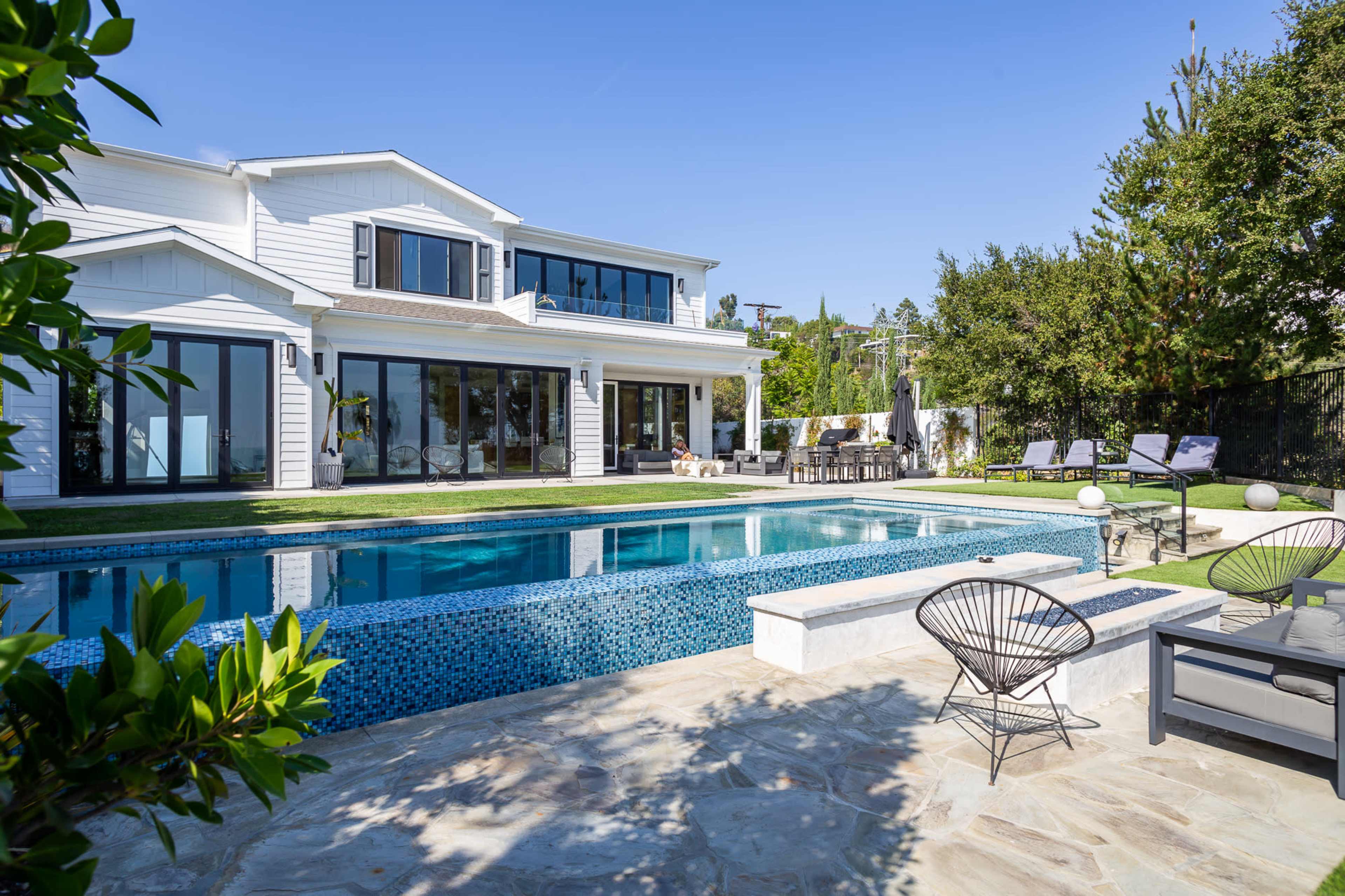 A modern house with large windows overlooks a swimming pool surrounded by lounge chairs and a patio area.