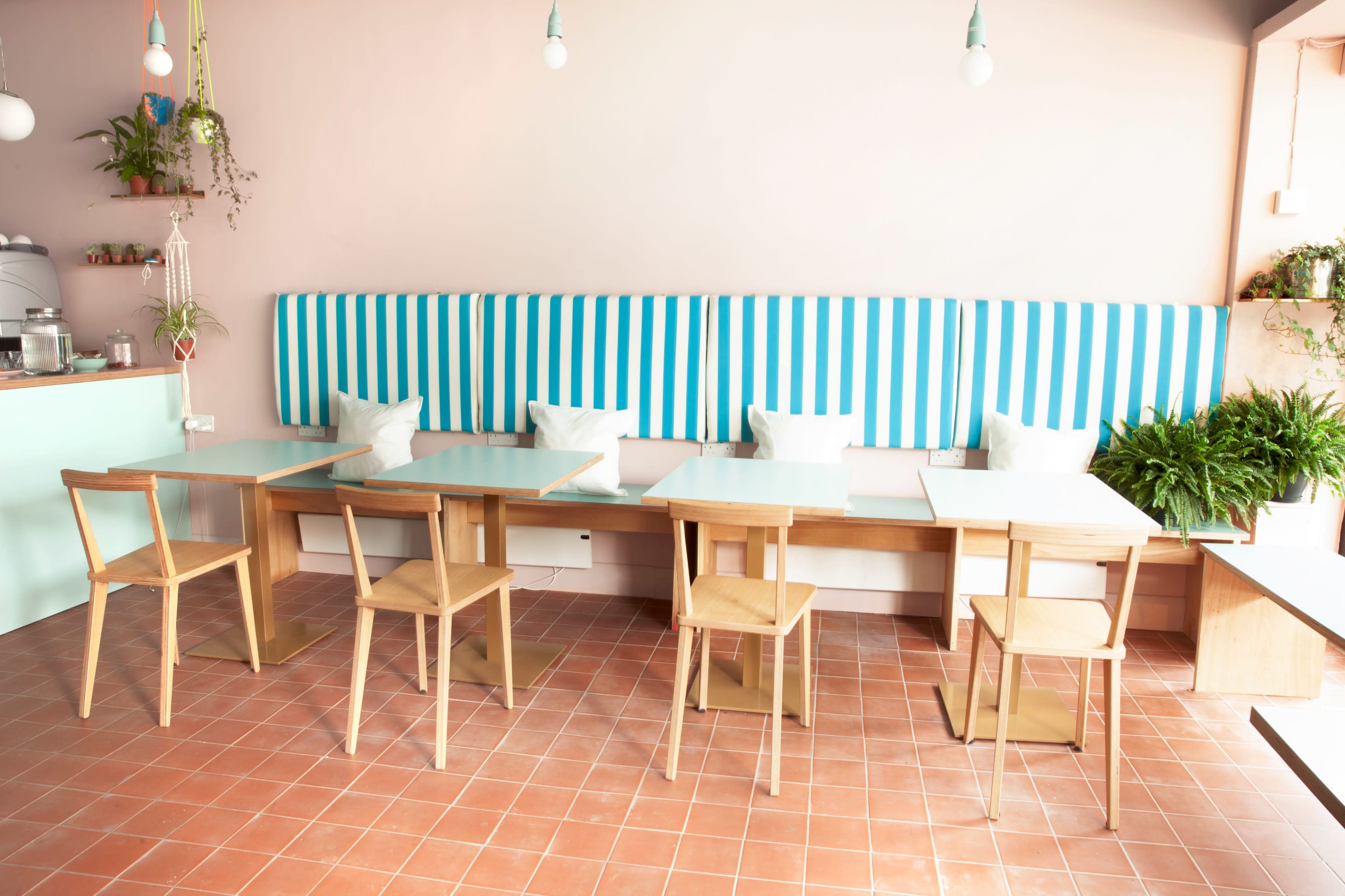 A modern cafe interior with light-colored walls, striped seating, wooden tables, and empty chairs arranged neatly.