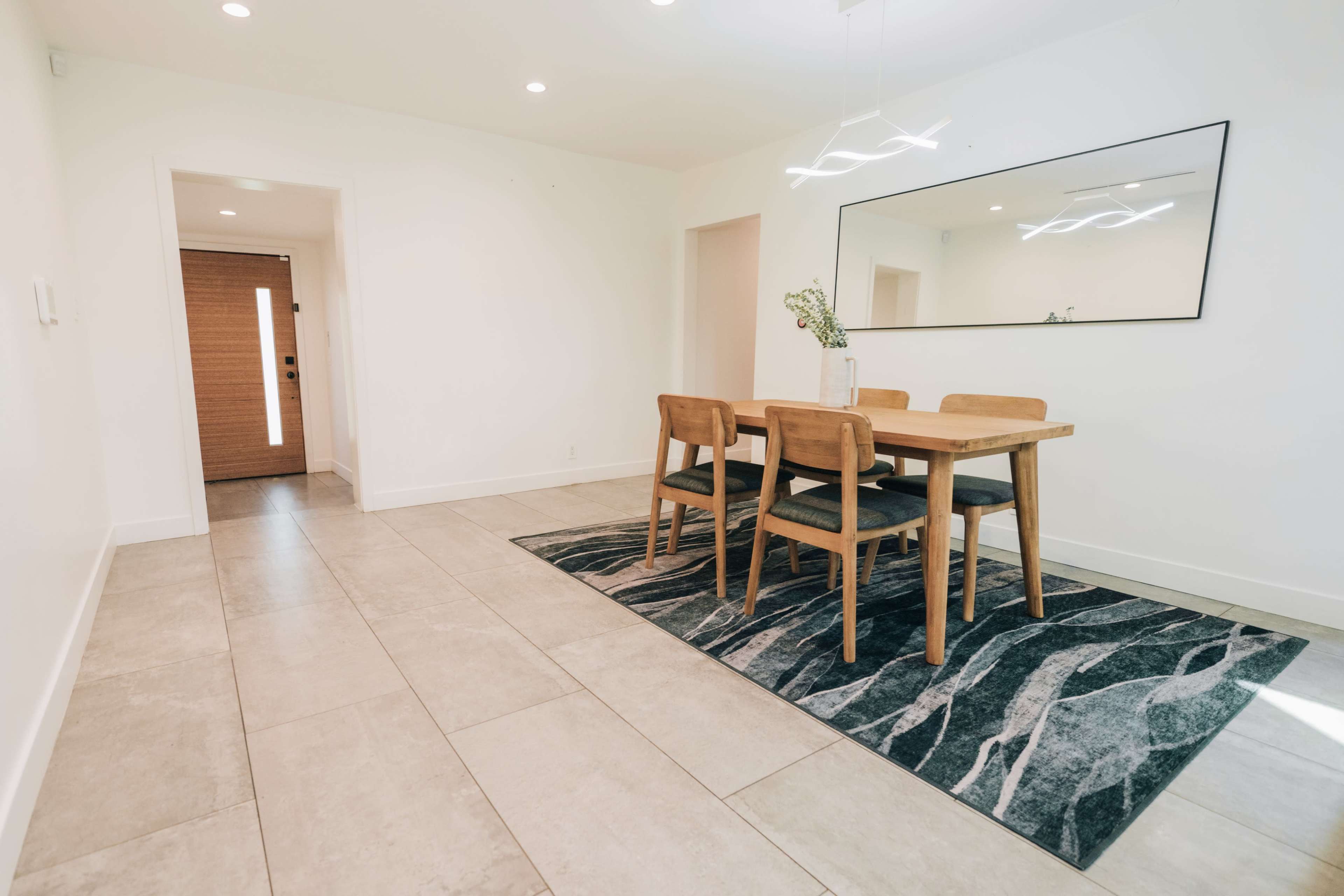 The image shows a dining area featuring a wooden table with four chairs, a large mirror on the wall, and a rug on tiled flooring, with a door in the background.