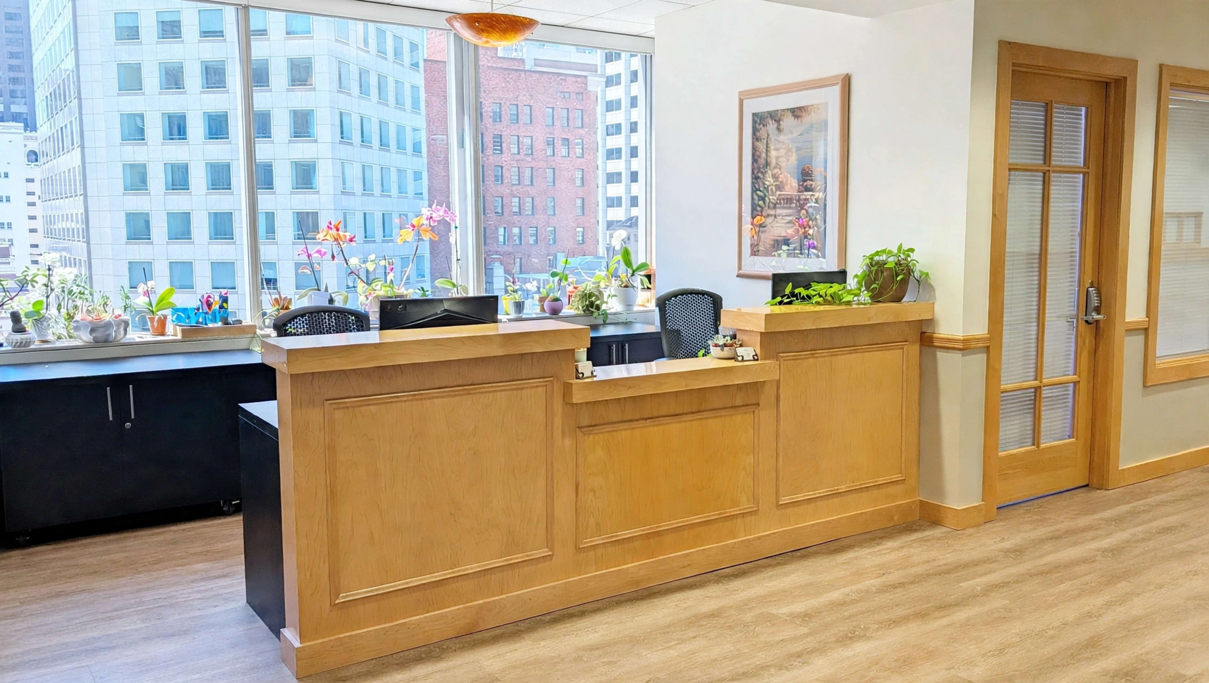 The image shows a reception area with a wooden front desk, potted plants on the counter, and large windows revealing a cityscape outside.