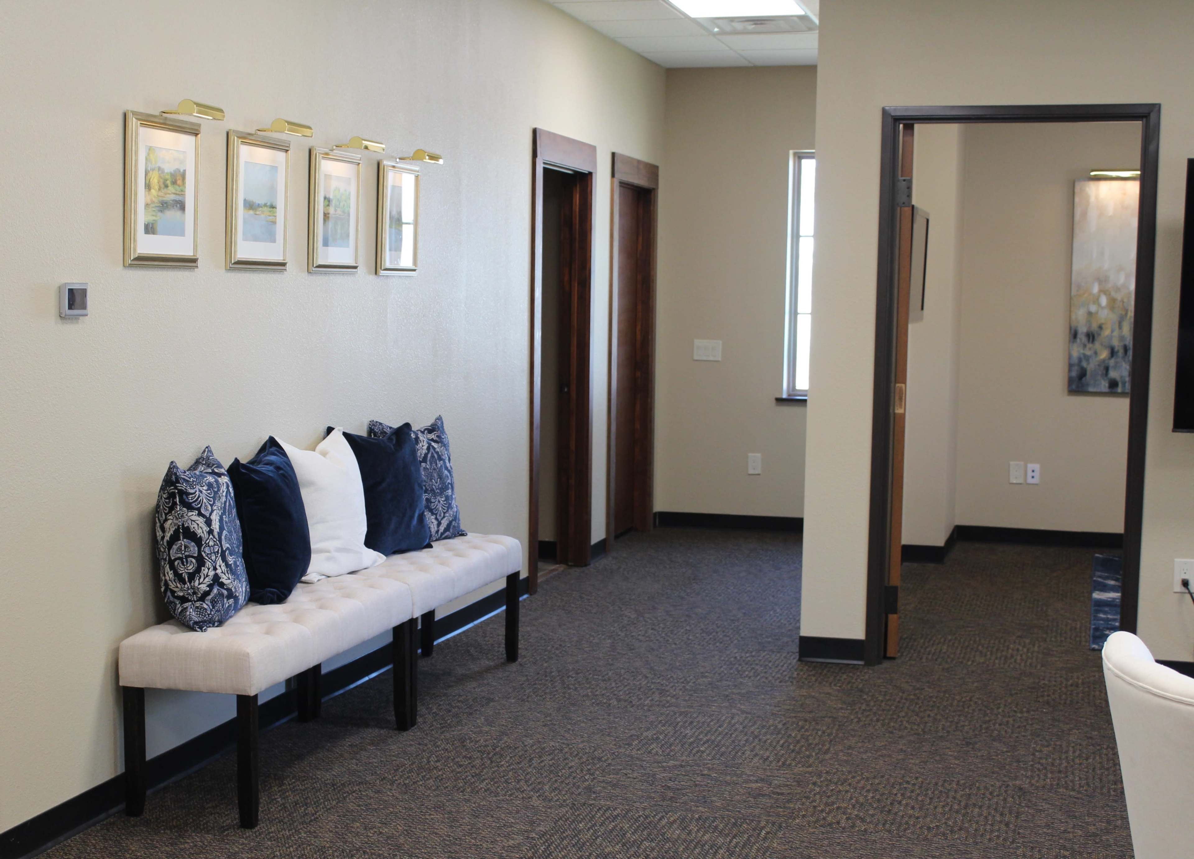 A well-lit hallway featuring a beige bench with blue and white pillows, framed artwork on the wall, and two closed doors opposite the bench.