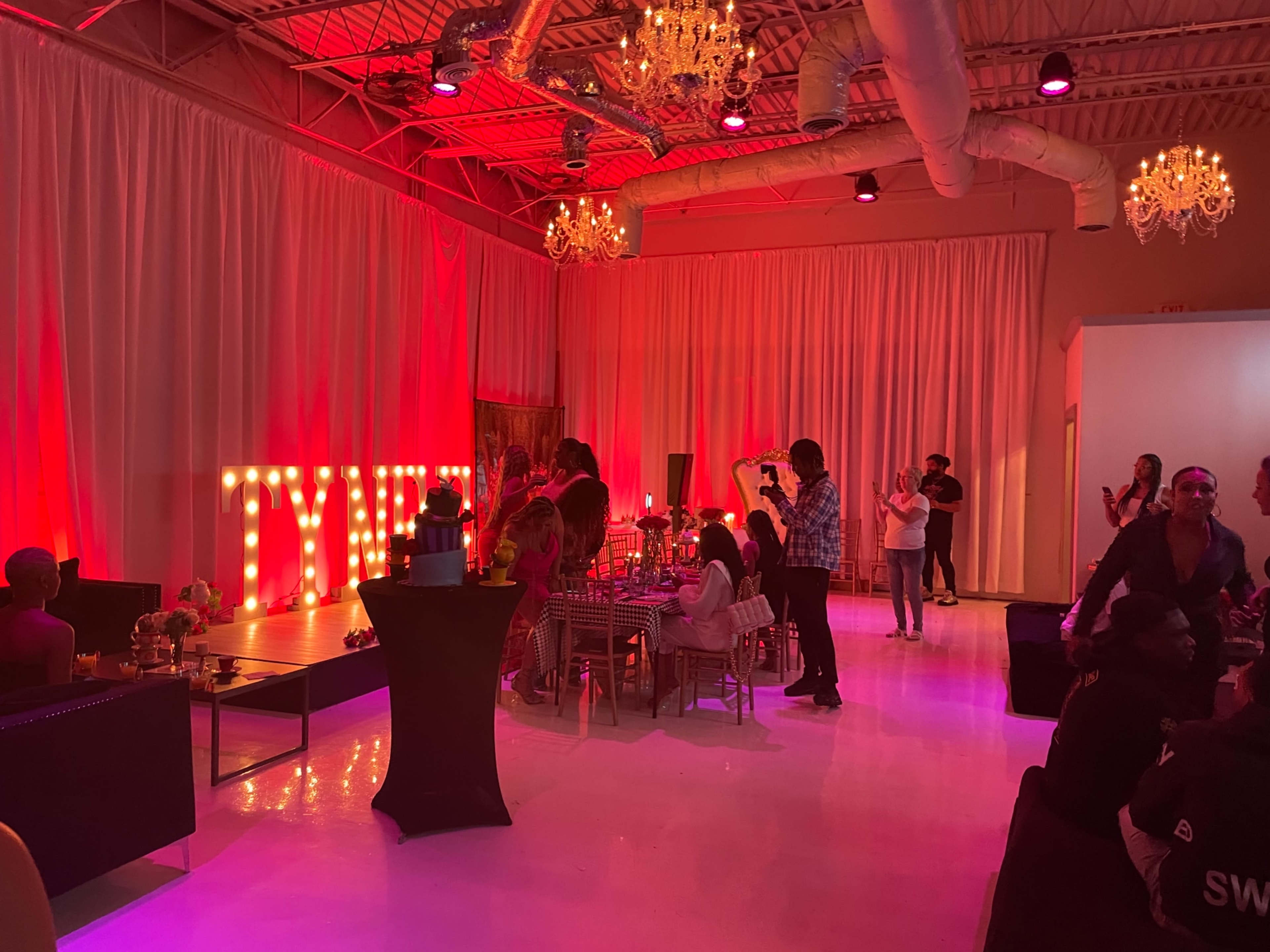 The scene shows a lively event space decorated with large illuminated letters spelling "TYME," featuring tables, guests mingling, and chandeliers hanging from the ceiling.