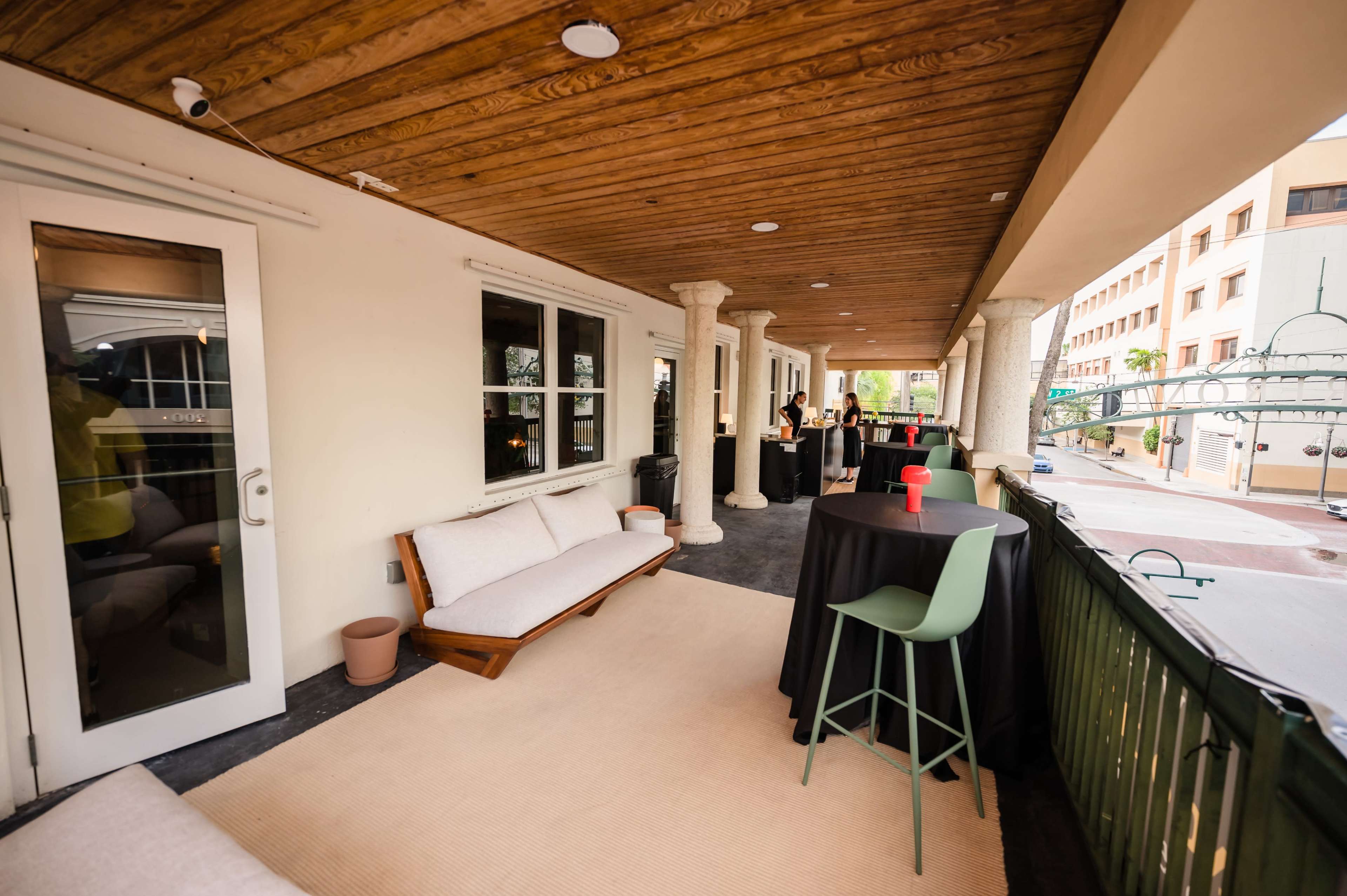A covered outdoor patio area featuring a couch, tall tables with chairs, and a view of a building exterior.