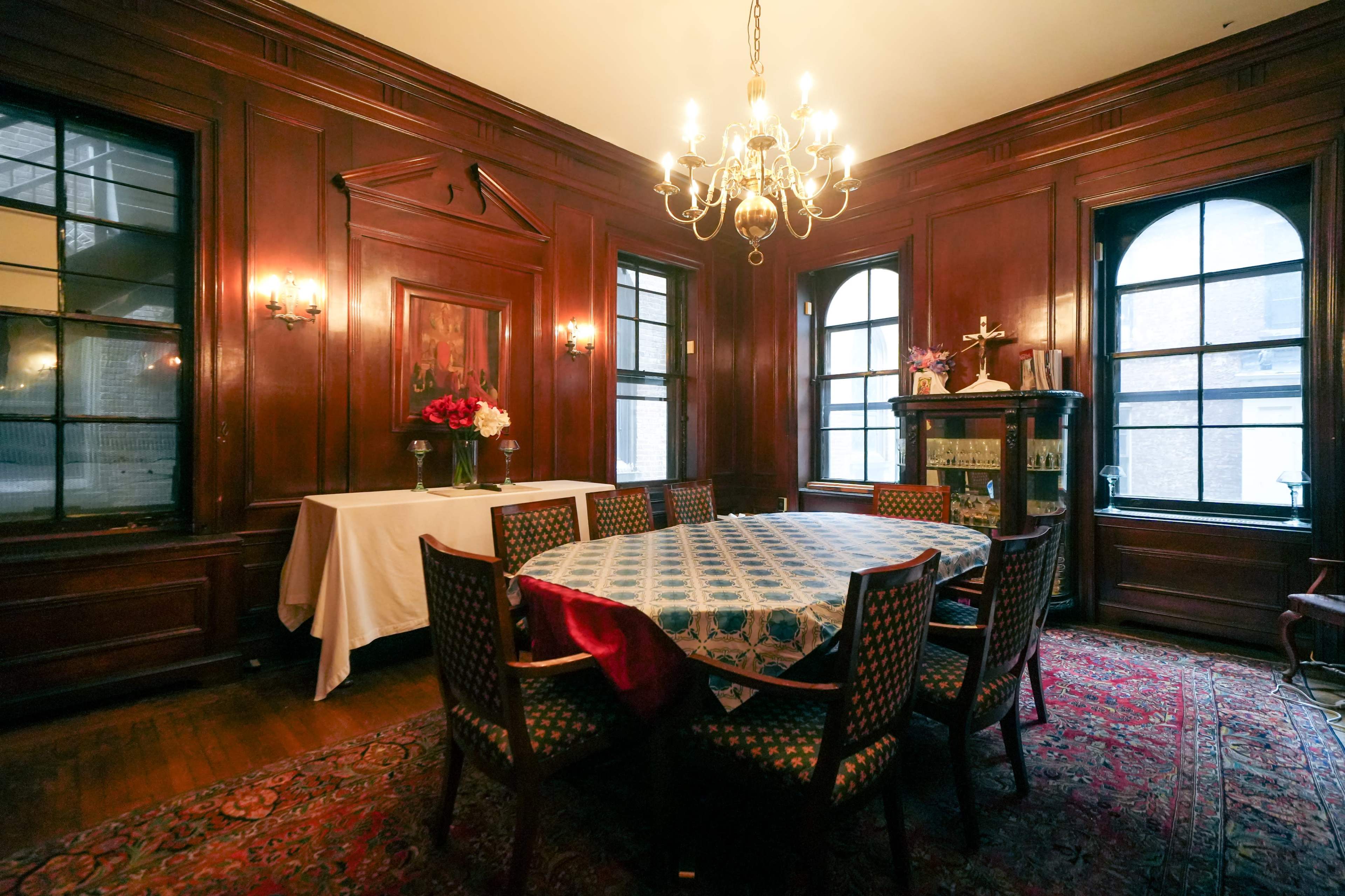 A dining room features a large table surrounded by chairs, with wood-paneled walls, a chandelier, and a windowed corner displaying a decorative cabinet.