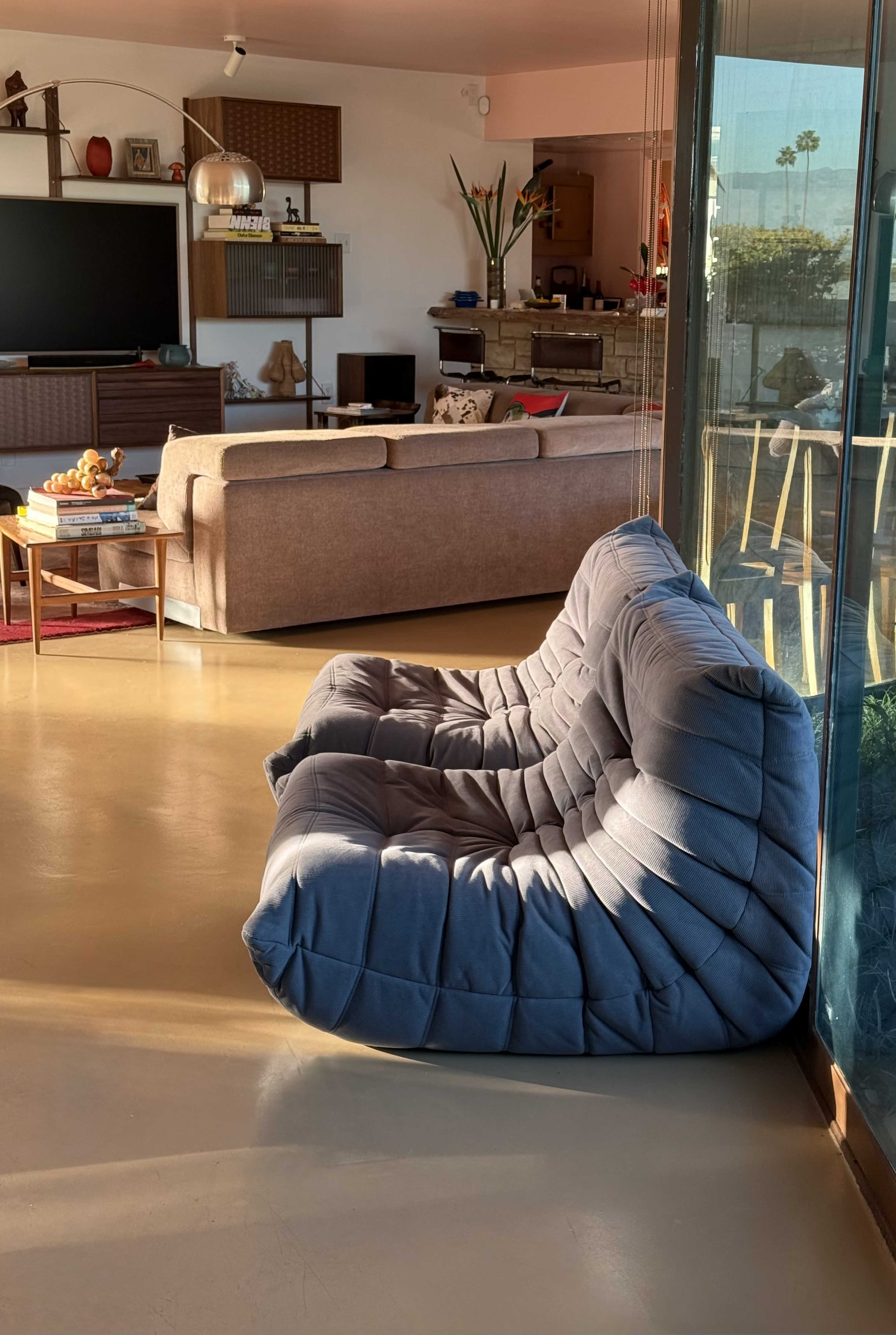 The image shows a modern living space featuring a gray, cushioned sofa positioned near large glass windows, with a view of a separate seating area and entertainment center.