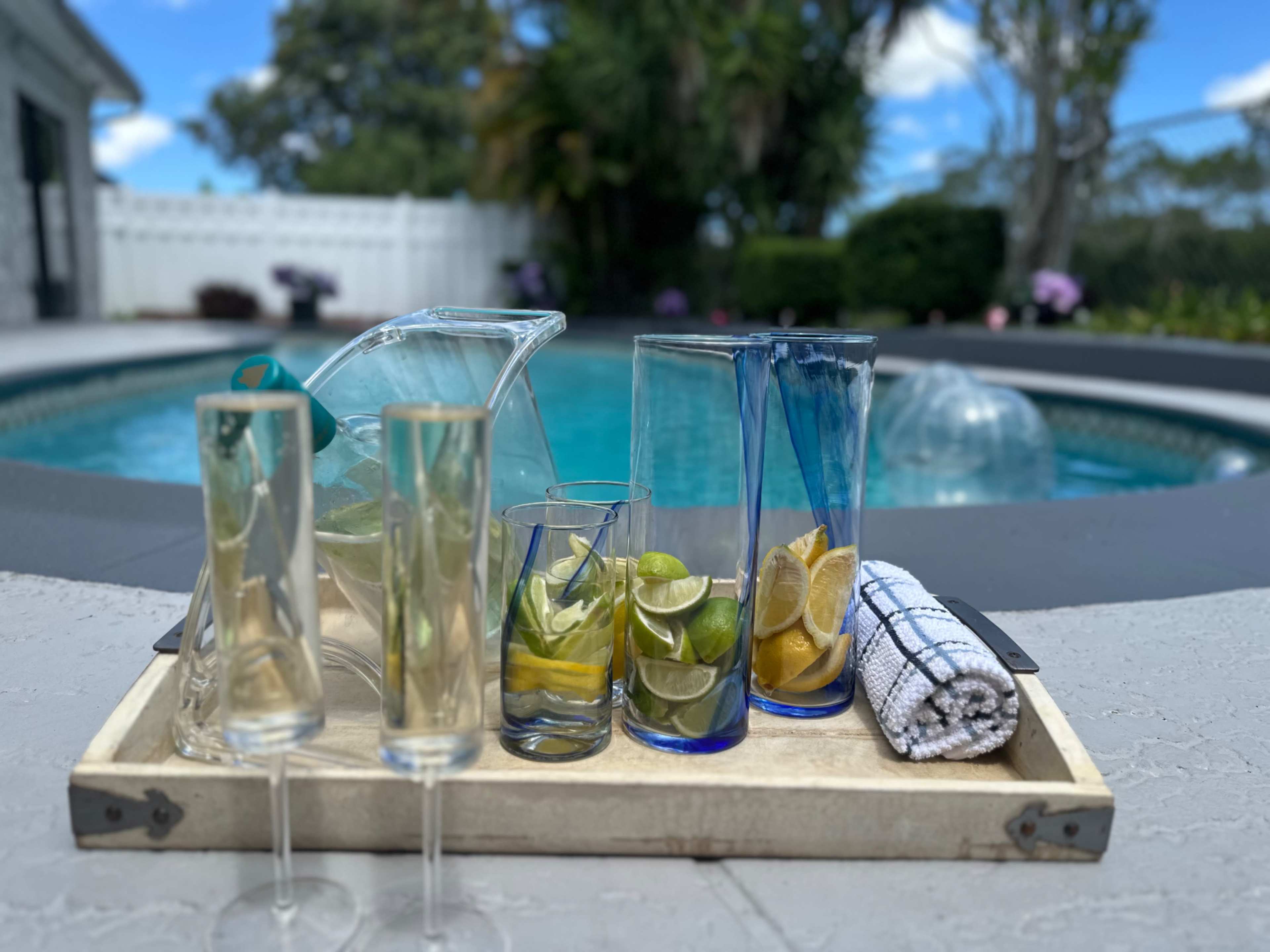 A wooden tray holds glasses filled with sliced citrus fruits and a pitcher near a swimming pool in a backyard setting.