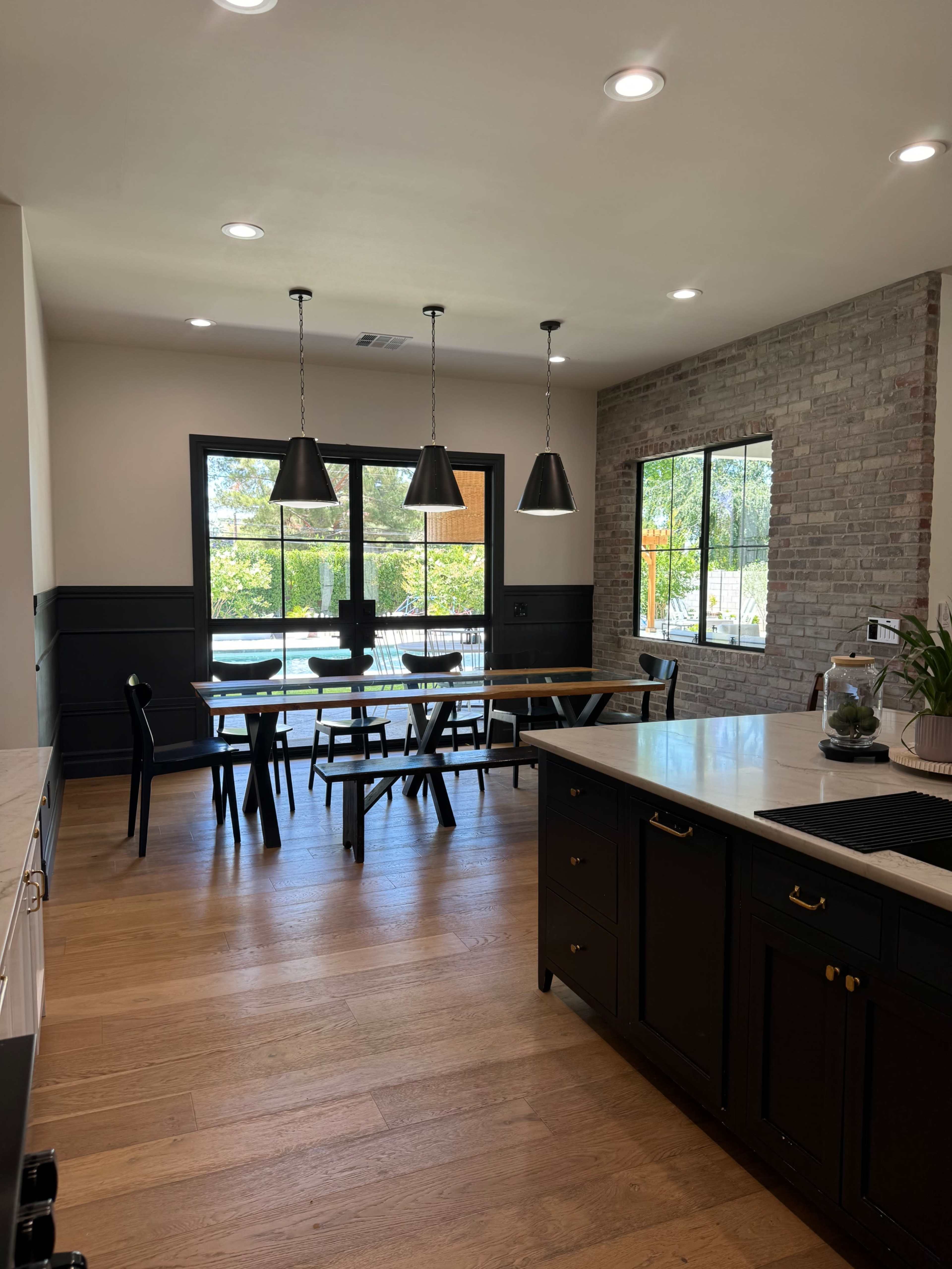 The kitchen features a dining area with a long table, black chairs, pendant lights, and large windows overlooking a garden.