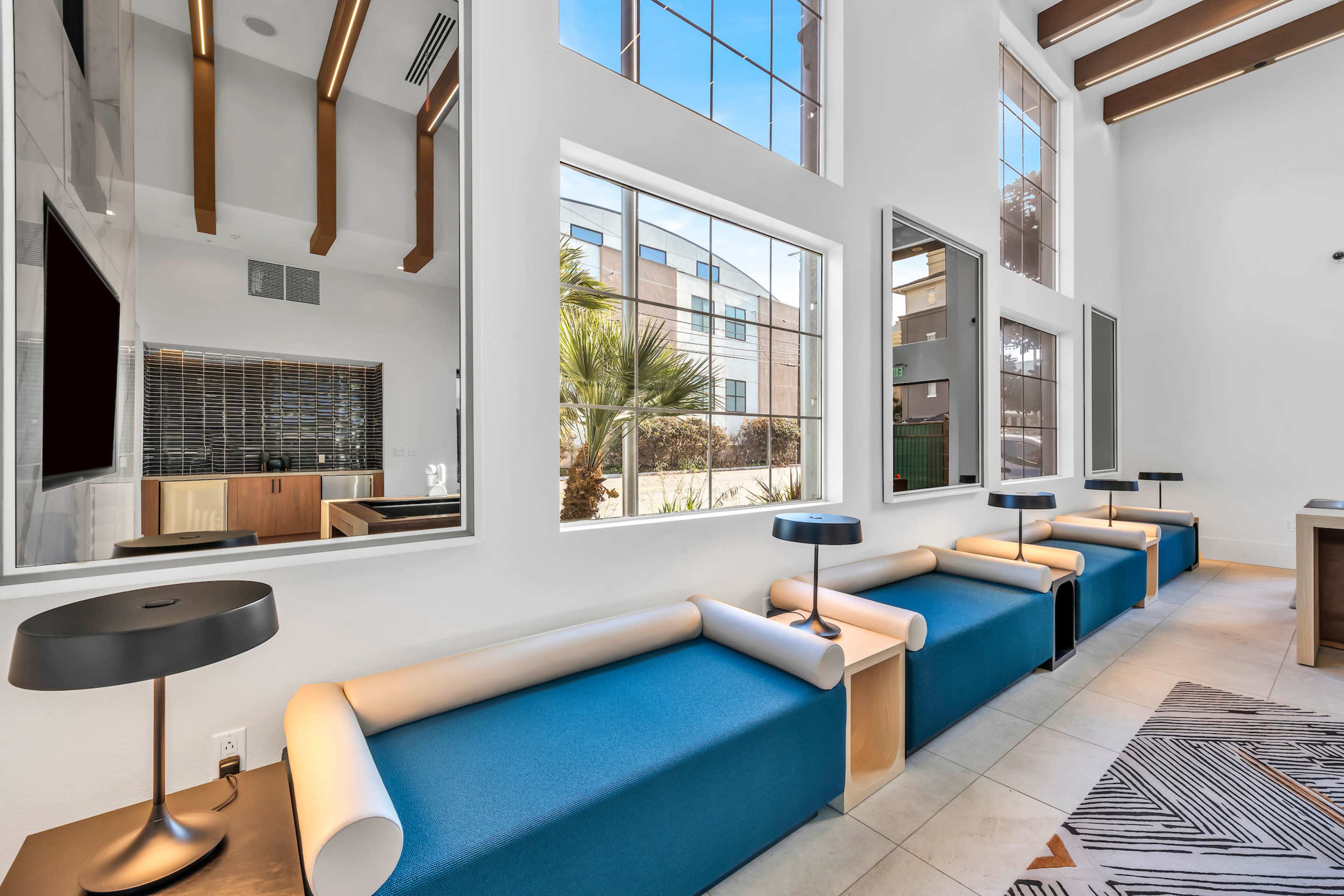 A modern interior space featuring blue seating with cylindrical armrests, large windows, and decorative lamps.