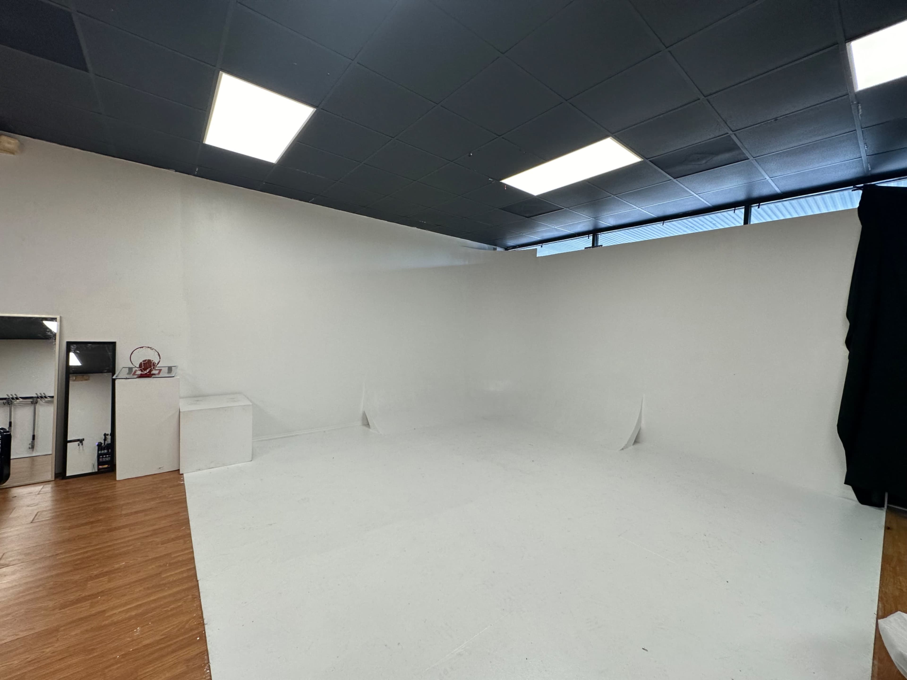 A spacious, empty studio with white walls and a light-colored floor, featuring a small area with equipment and a black curtain in one corner.