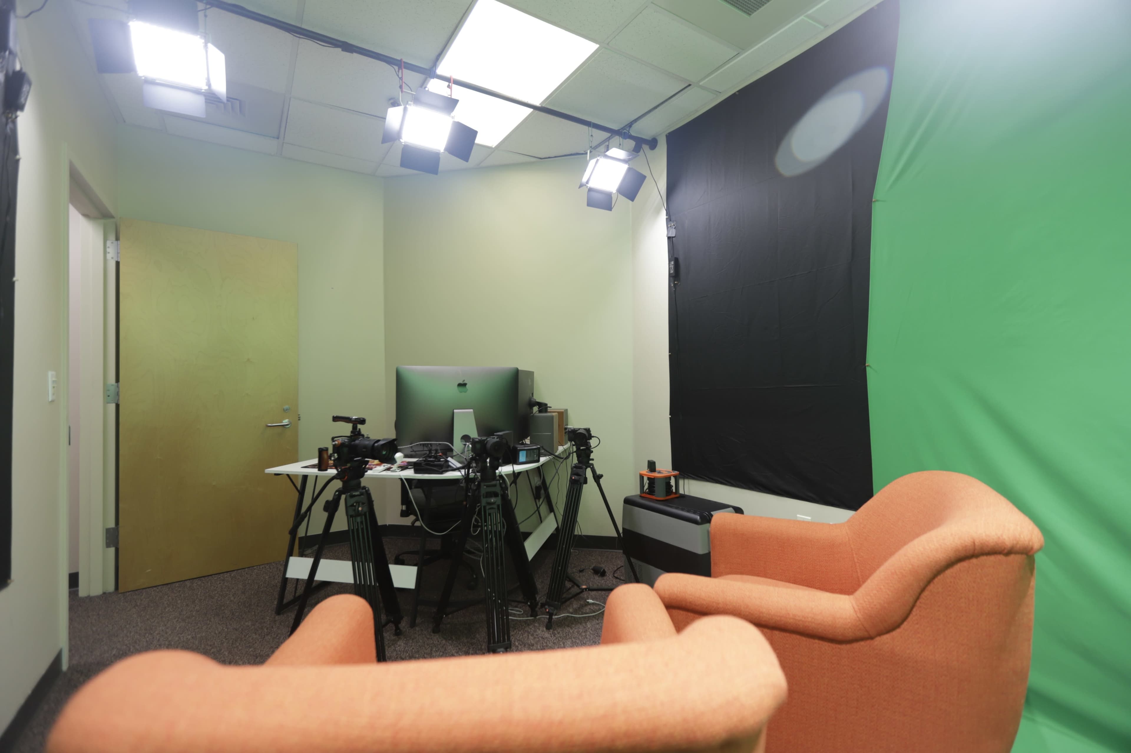 A studio setup featuring two orange chairs, a green screen, and various camera equipment arranged around a central desk with a computer.