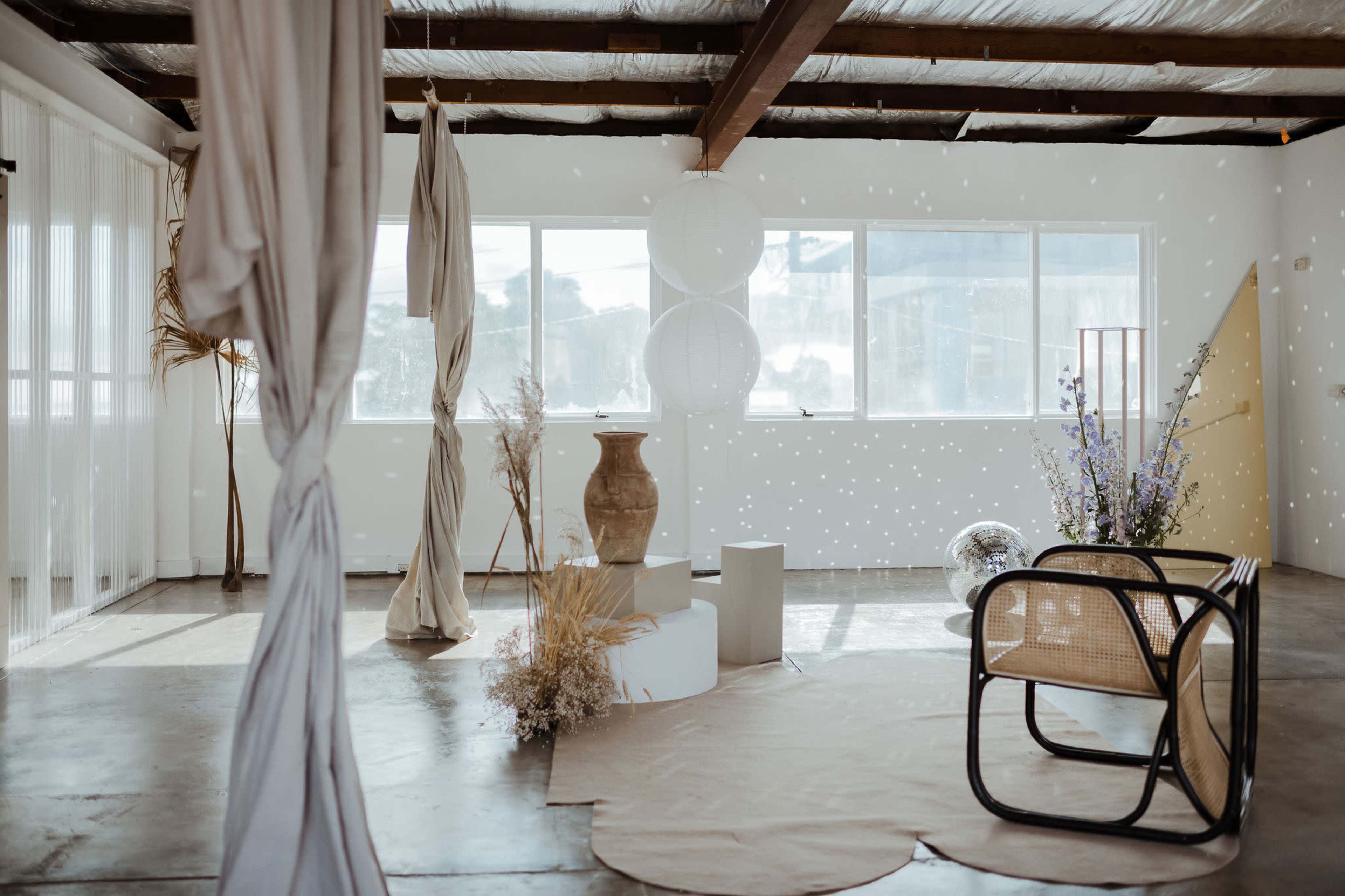 A spacious, minimalist room with large windows, featuring a decorative arrangement of a clay pot, plants, and various textures including fabric drapes and a rattan chair.