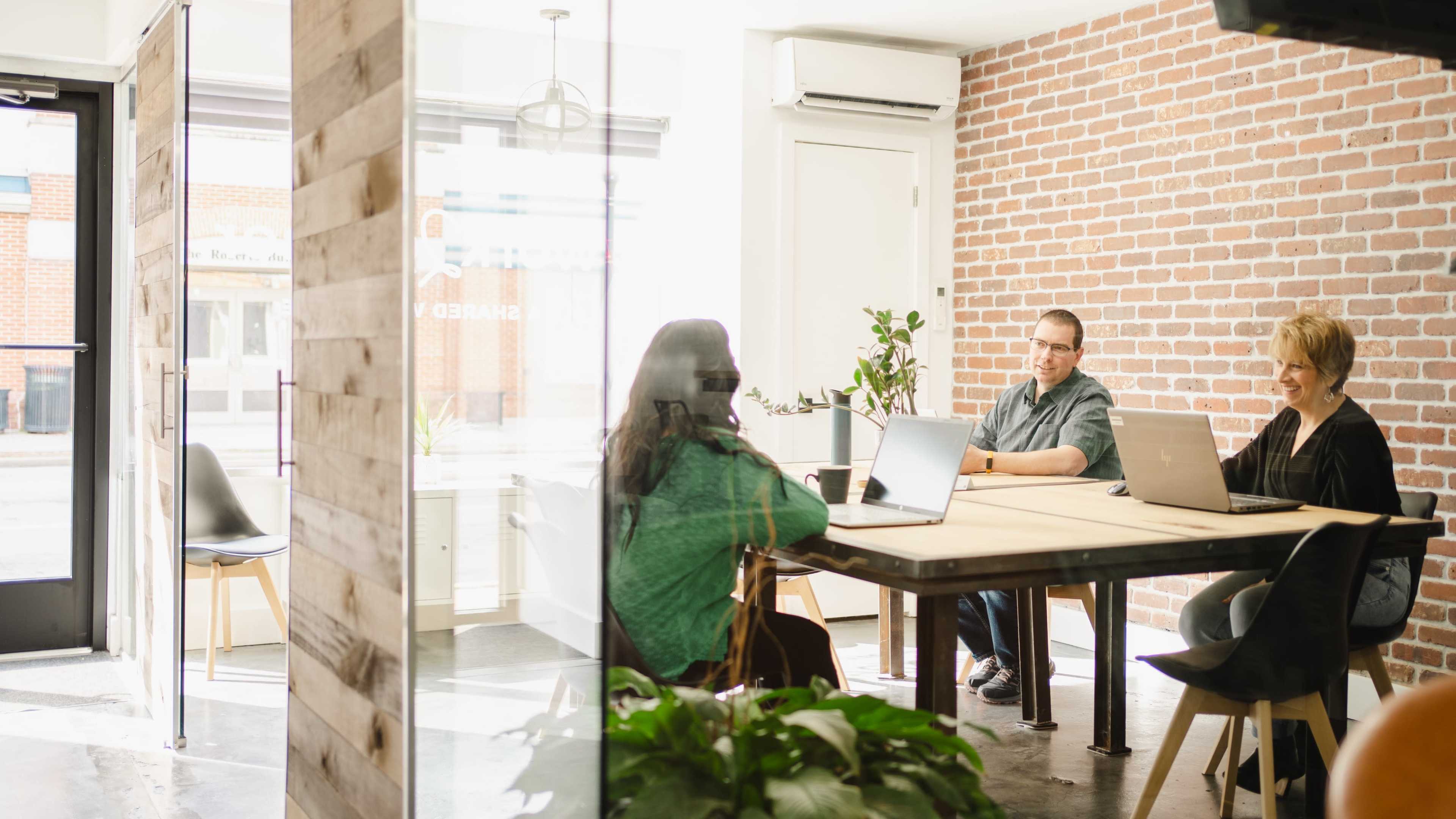 Three people are sitting around a table in a modern office space with large windows and exposed brick walls, each working on laptops.
