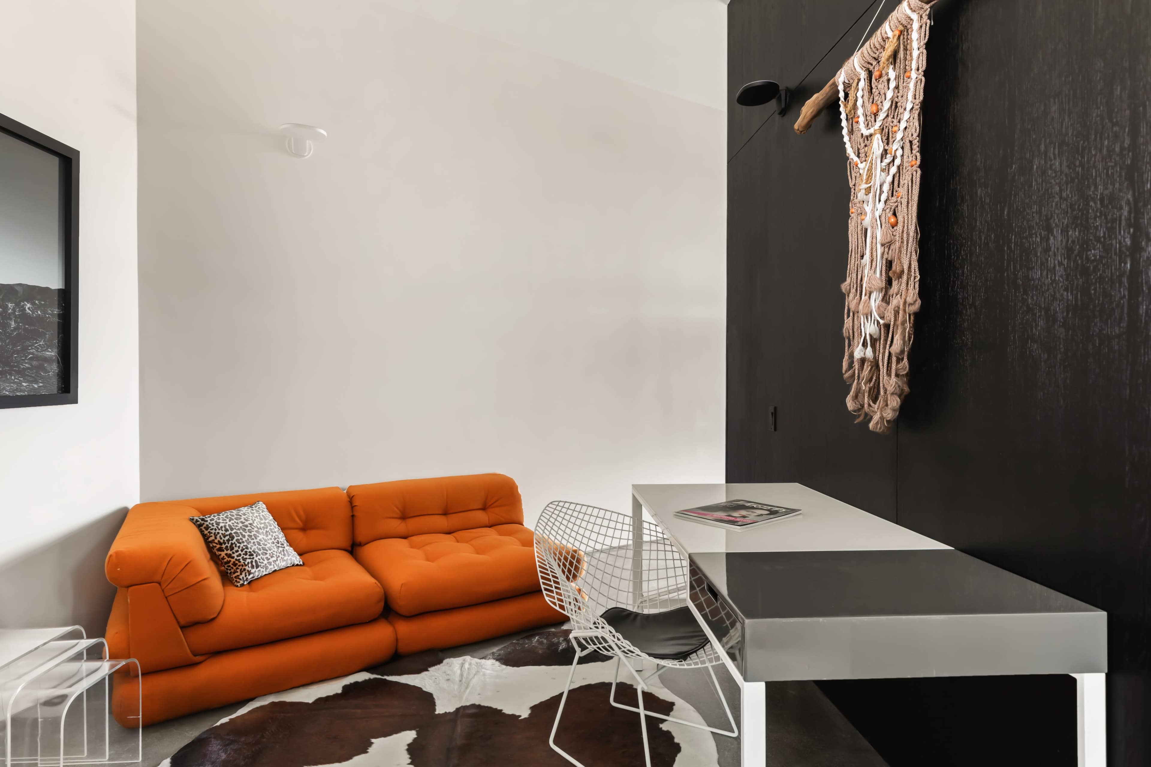 The image features a modern interior with an orange sofa, a white desk with a wire chair, and a wall adorned with a textile hanging.