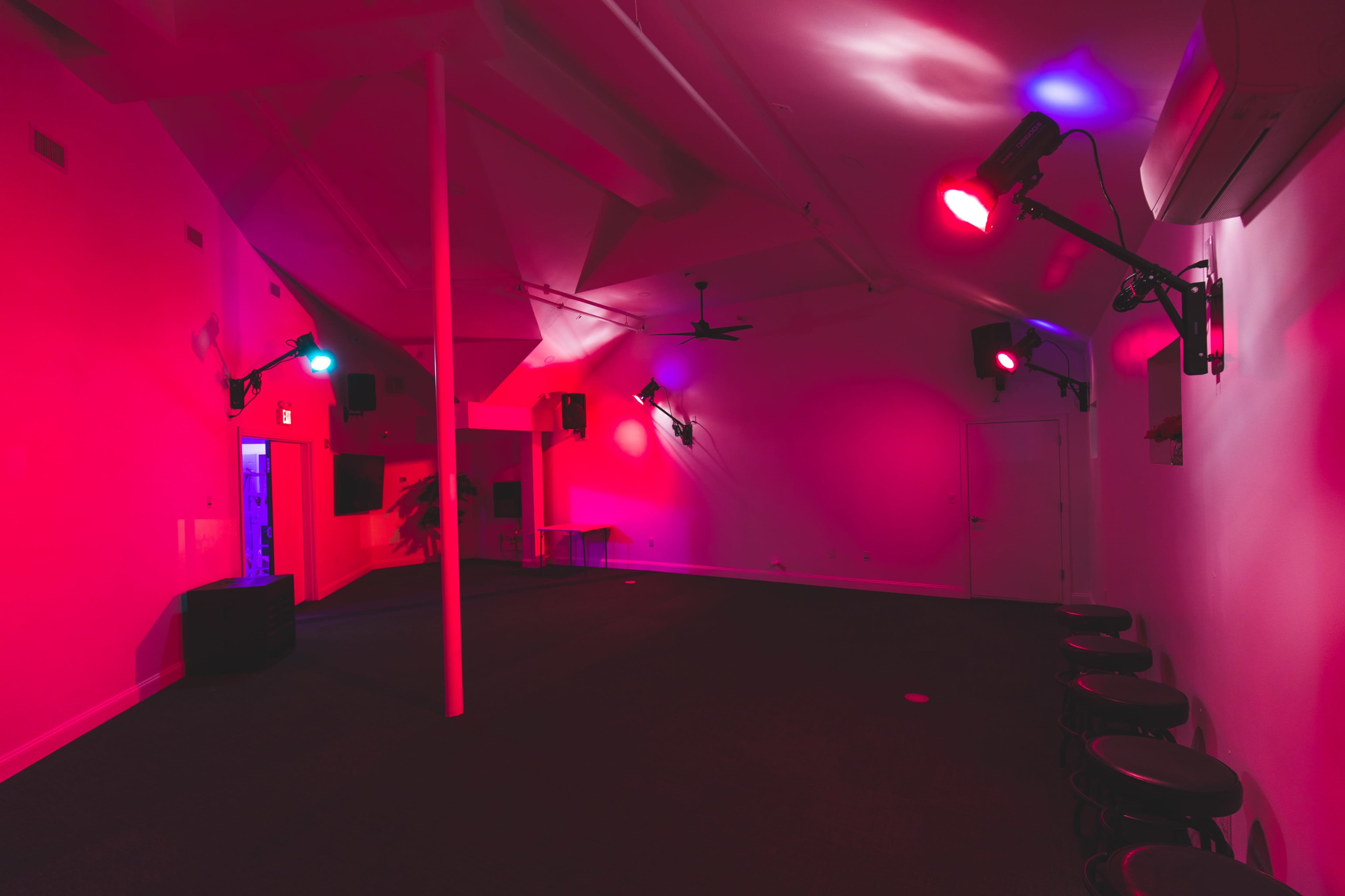 A dimly lit room with pink and blue lighting, featuring several wall-mounted lights and a few bar stools along one side.