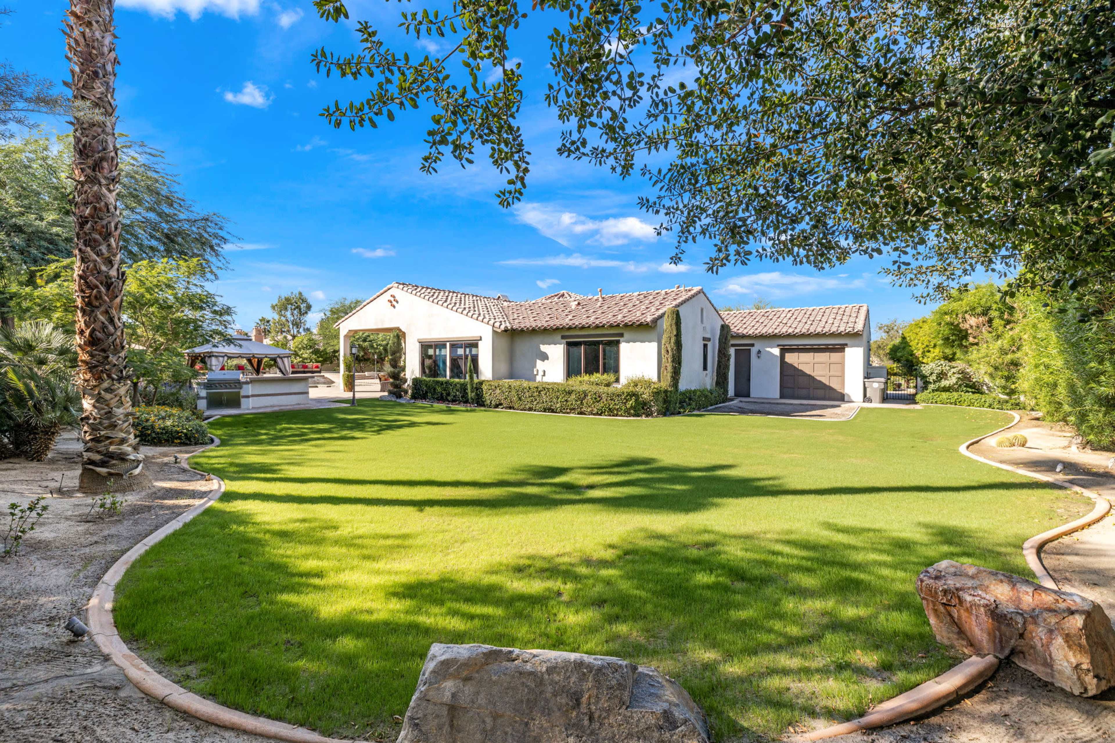 A single-story house with a tiled roof sits beside a neatly landscaped lawn and garden, featuring palm trees and a small patio area.