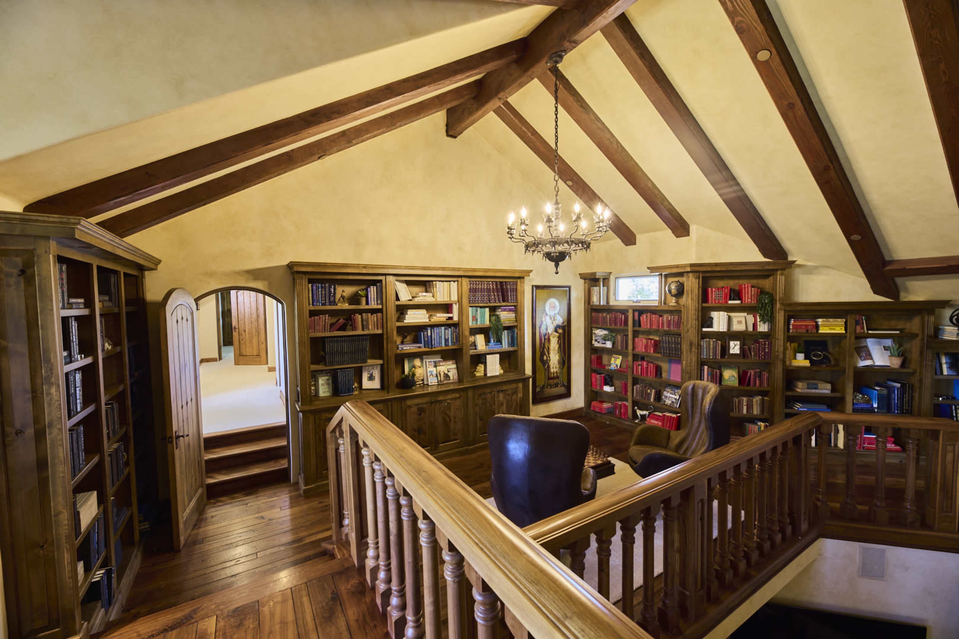 A spacious loft area features wooden beams, a chandelier, and shelves filled with books surrounding a pair of leather chairs.