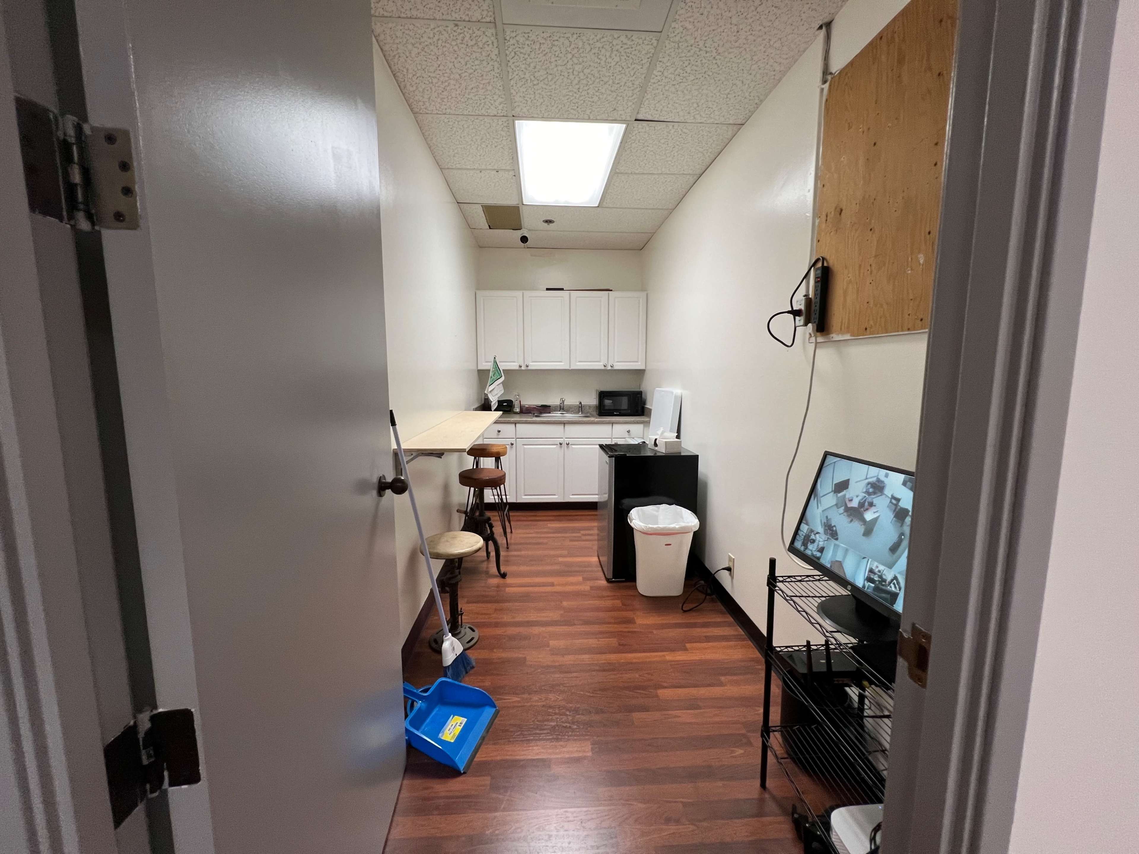 A narrow room with a kitchenette on one side, a small table with two stools, a trash can, and a TV displaying security footage.