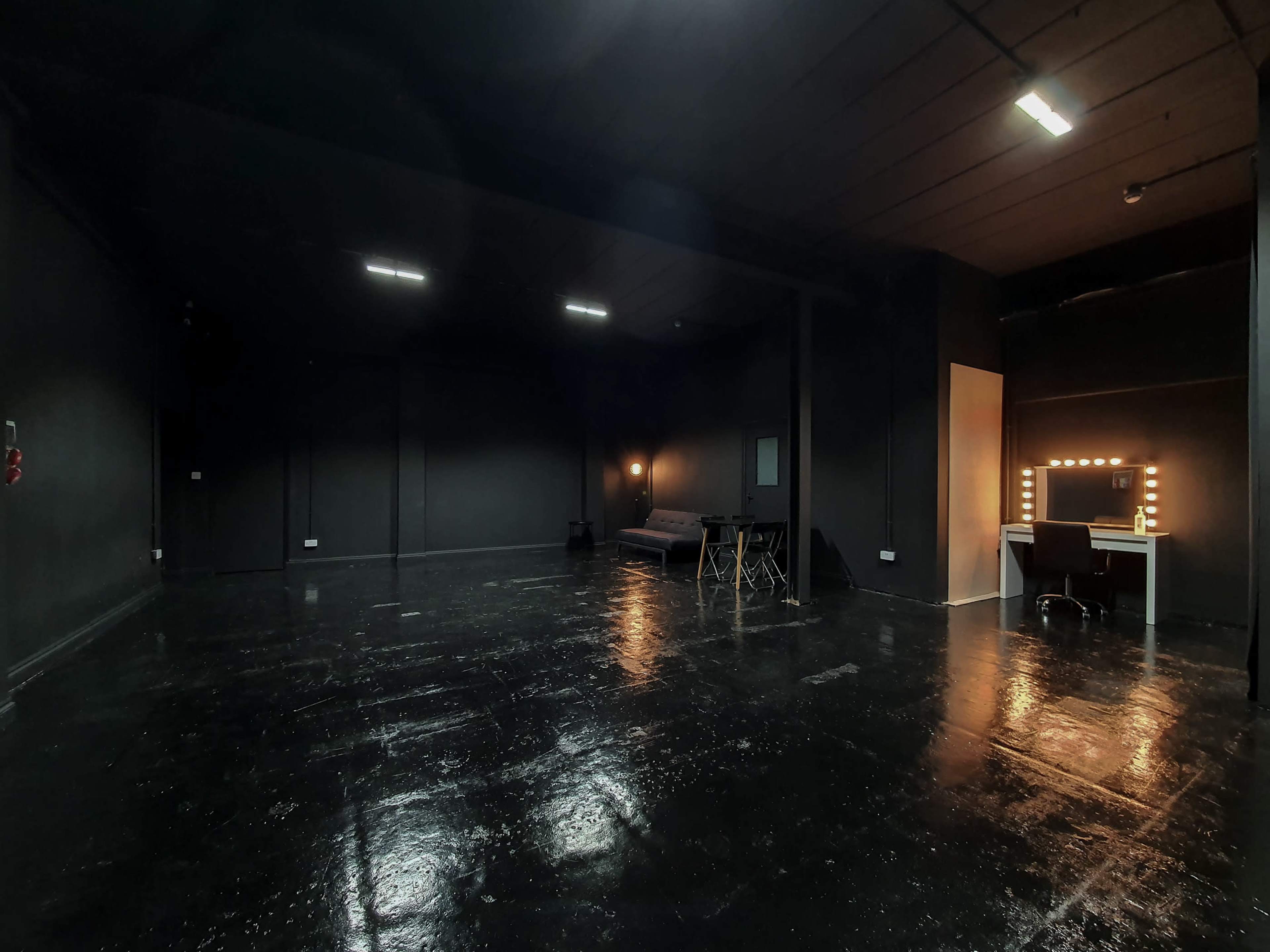 A large, empty room with black walls and a polished black floor, featuring a vanity mirror with lights and a couch along one side.