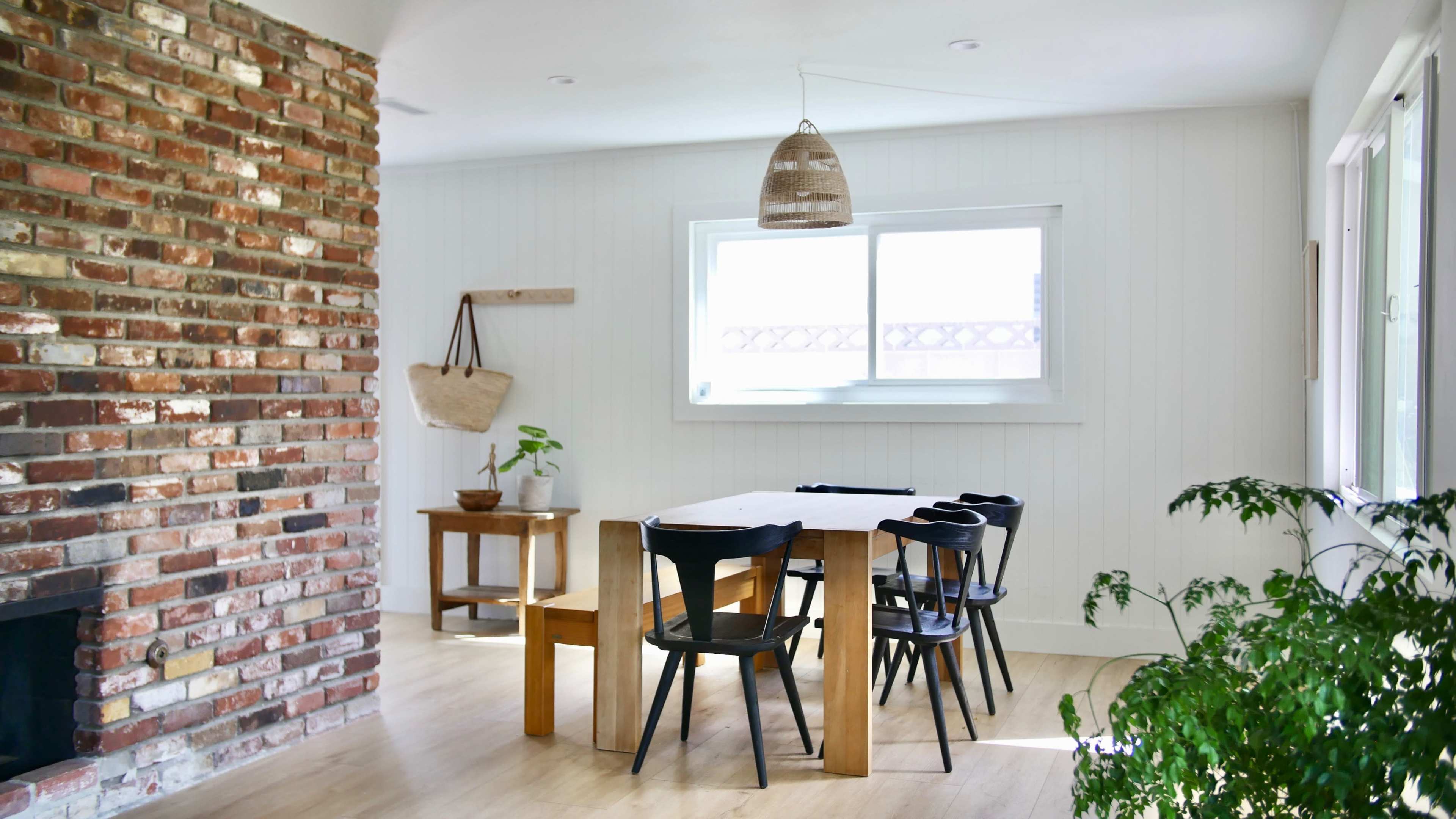 A dining area with a wooden table surrounded by black chairs, a brick wall, and a large window letting in natural light.