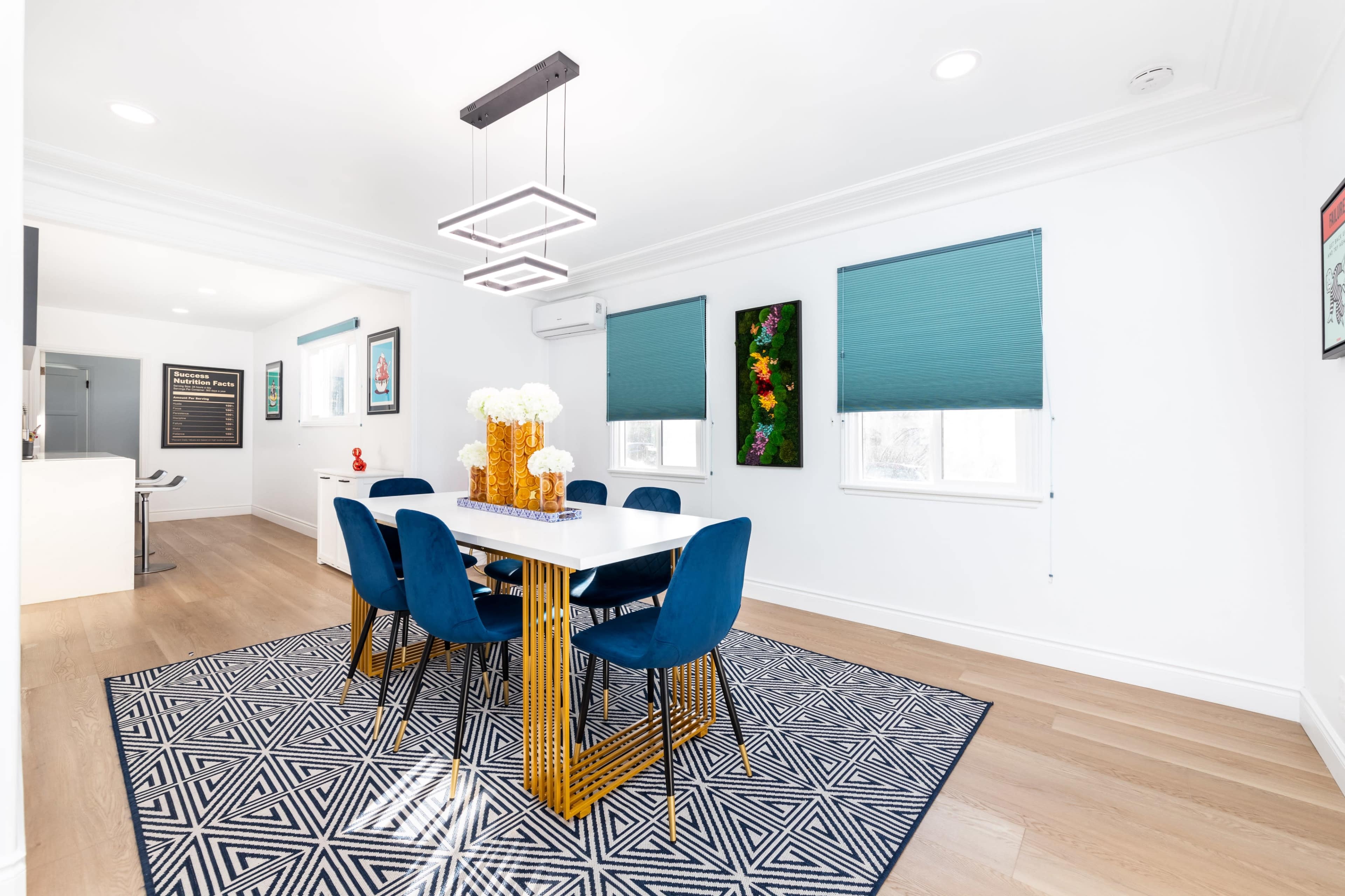 A modern dining area features a white table with blue chairs, a geometric patterned rug, and a light fixture overhead.