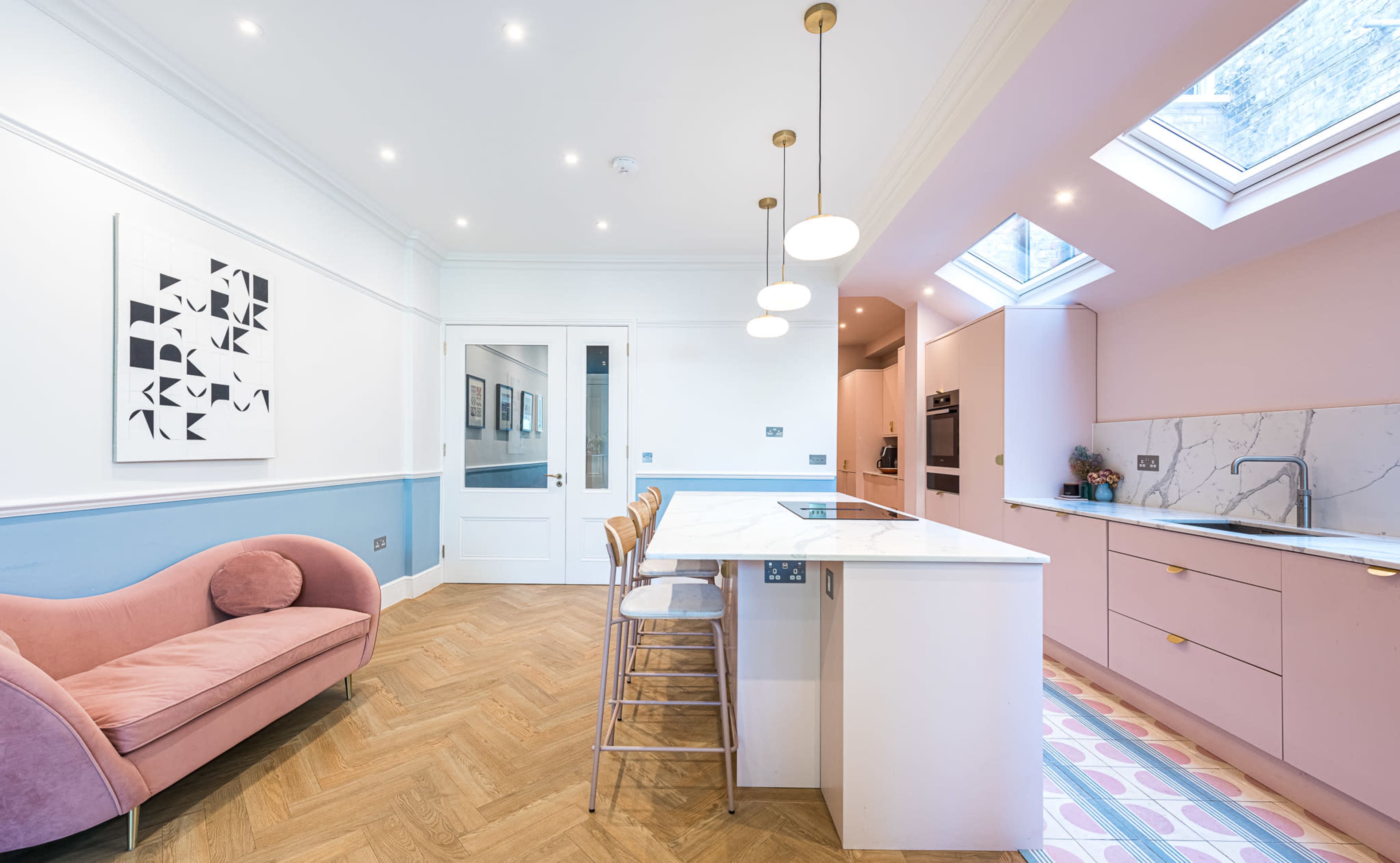 The image shows a modern kitchen with pink cabinetry, a marble island, a pink sofa, and skylights illuminating the space.