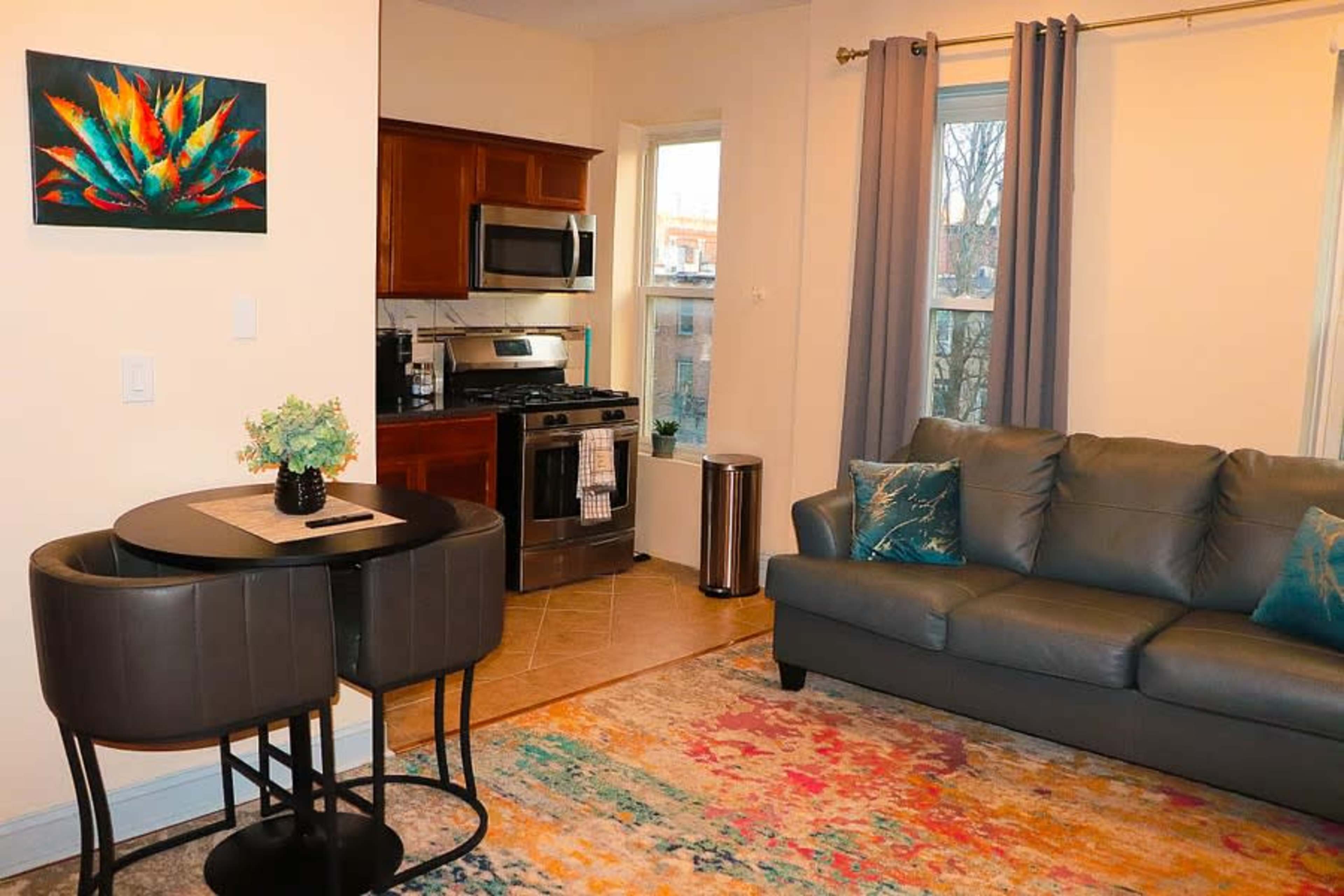 The image shows a living space featuring a gray couch, a small round dining table with two chairs, and a kitchen area with wooden cabinets and stainless steel appliances.