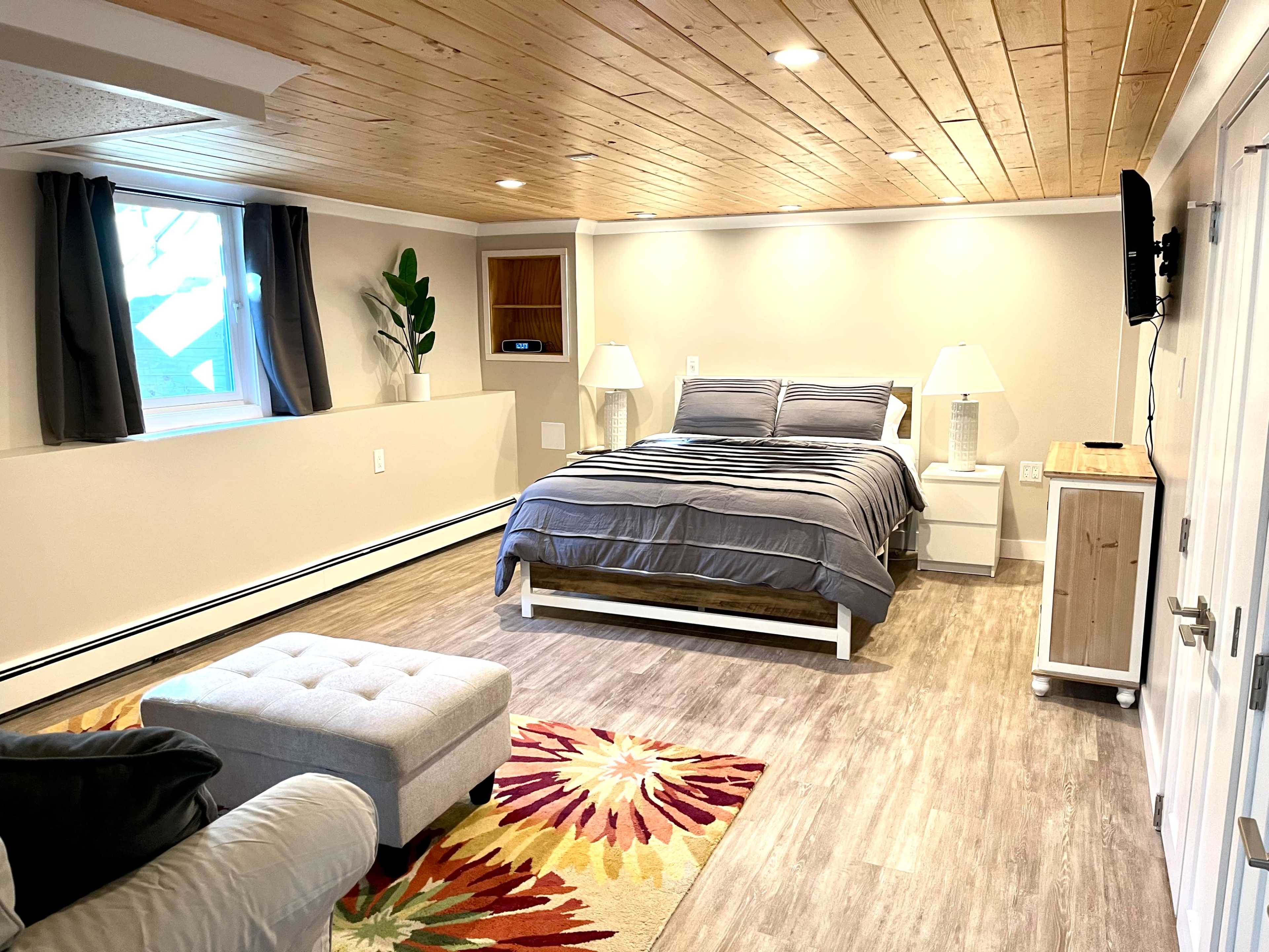A spacious, well-lit bedroom features a queen-sized bed, two nightstands with lamps, a plush ottoman, and a large window.