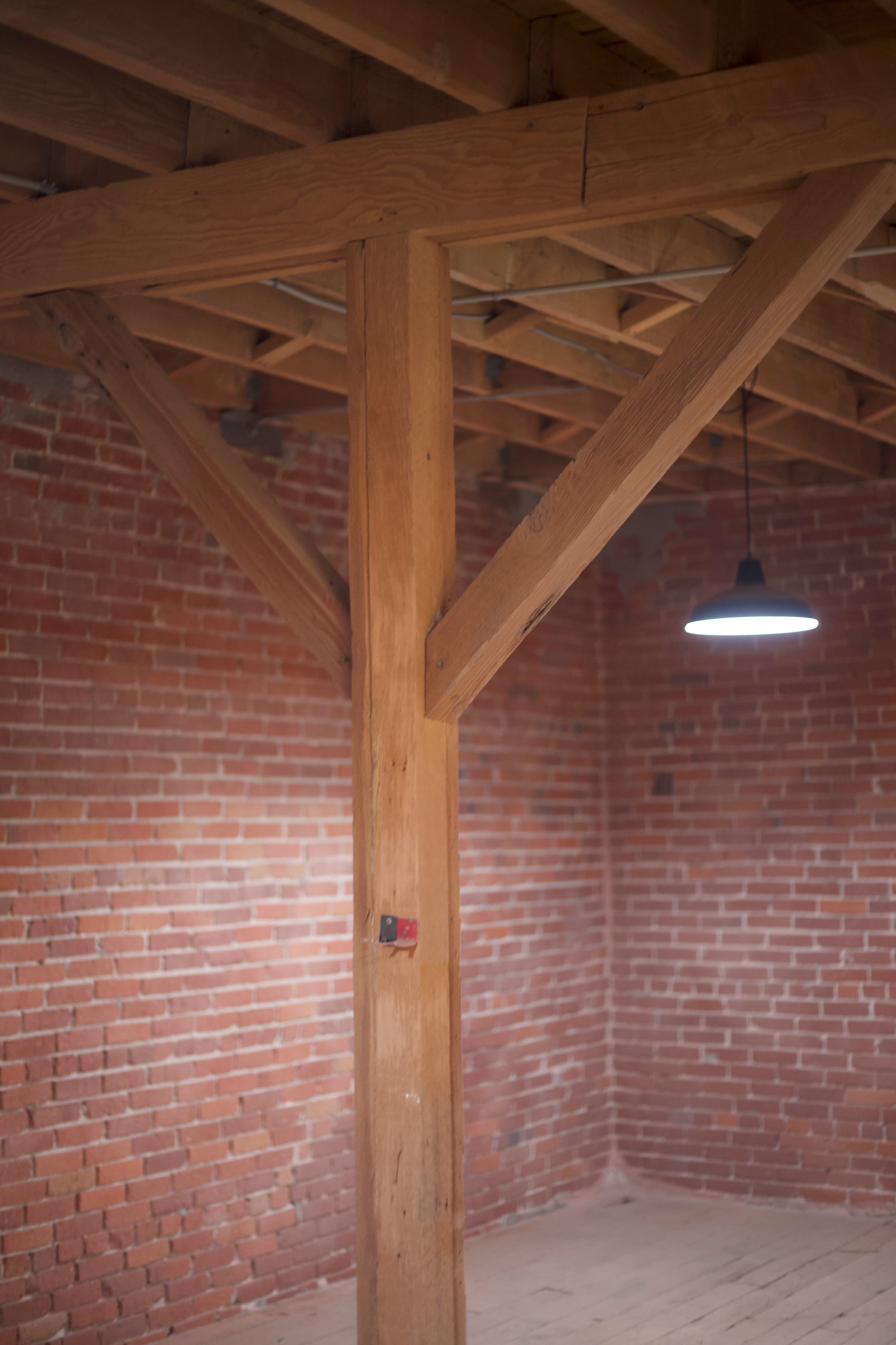 A bare room with exposed wooden beams and a single hanging light fixture, surrounded by red brick walls.