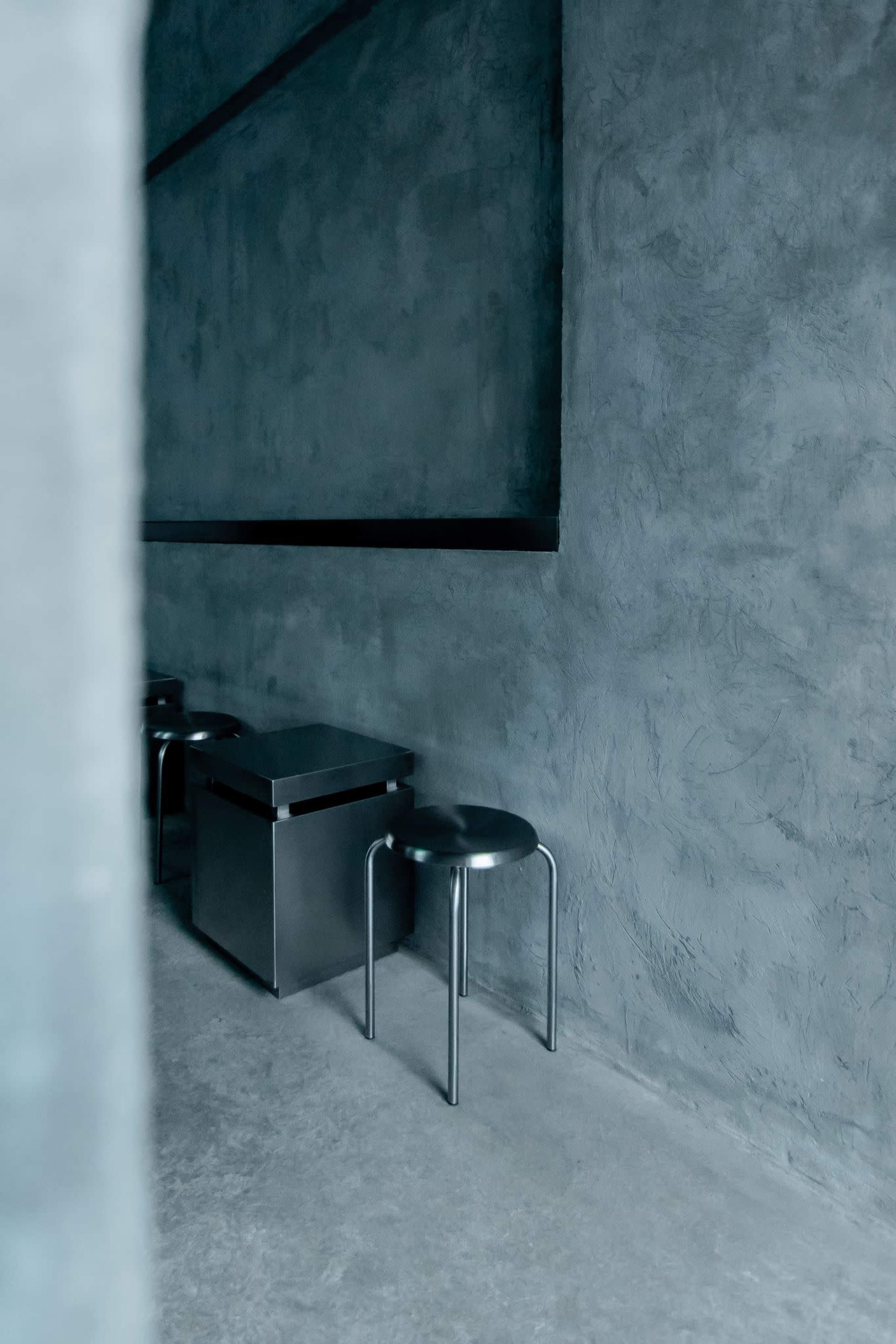 The image shows a minimalist interior with a concrete wall, a metal stool, and a small table.